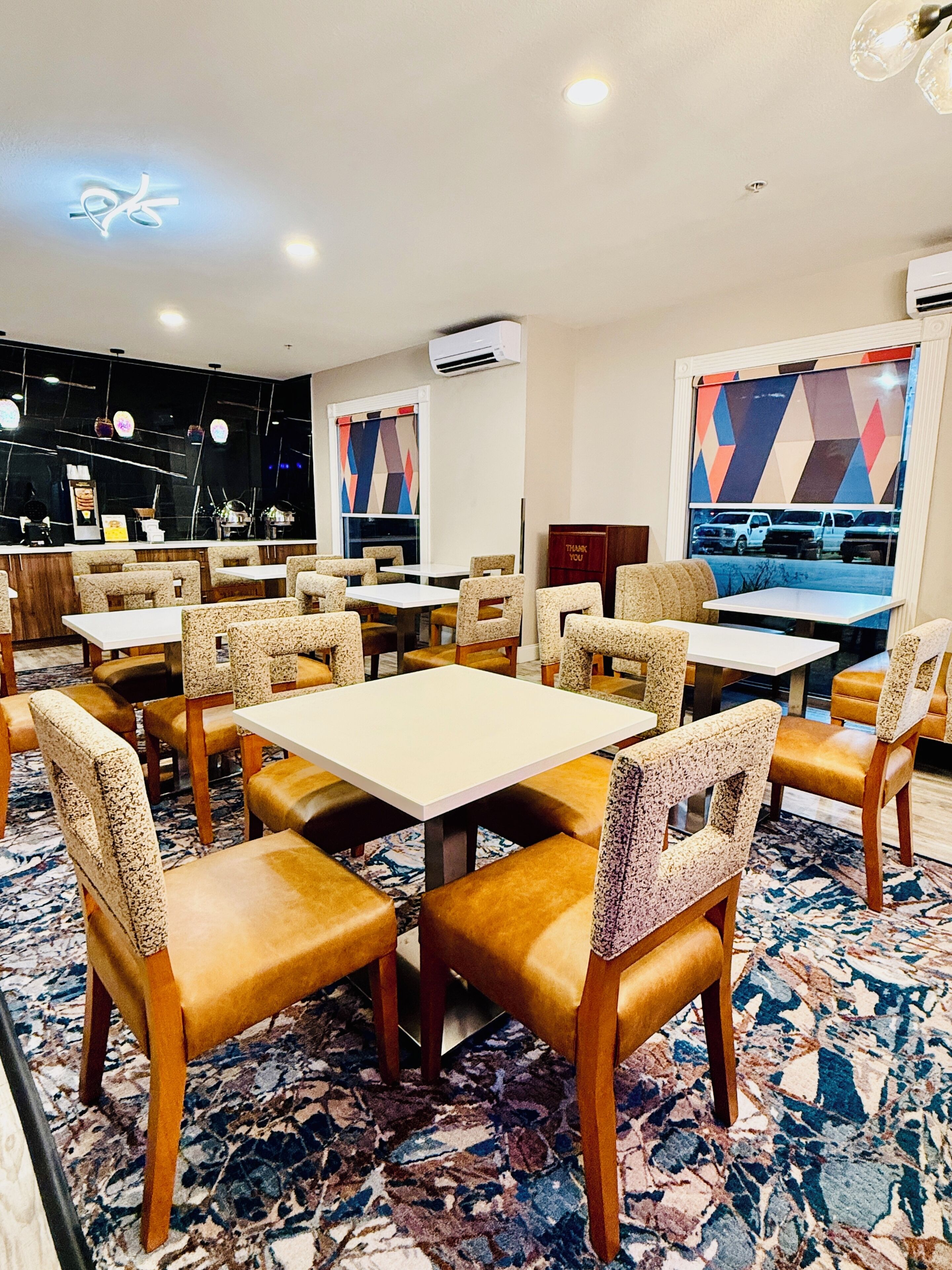 A restaurant interior with multiple tables and chairs, a patterned carpet, and a view of the outside through a window.