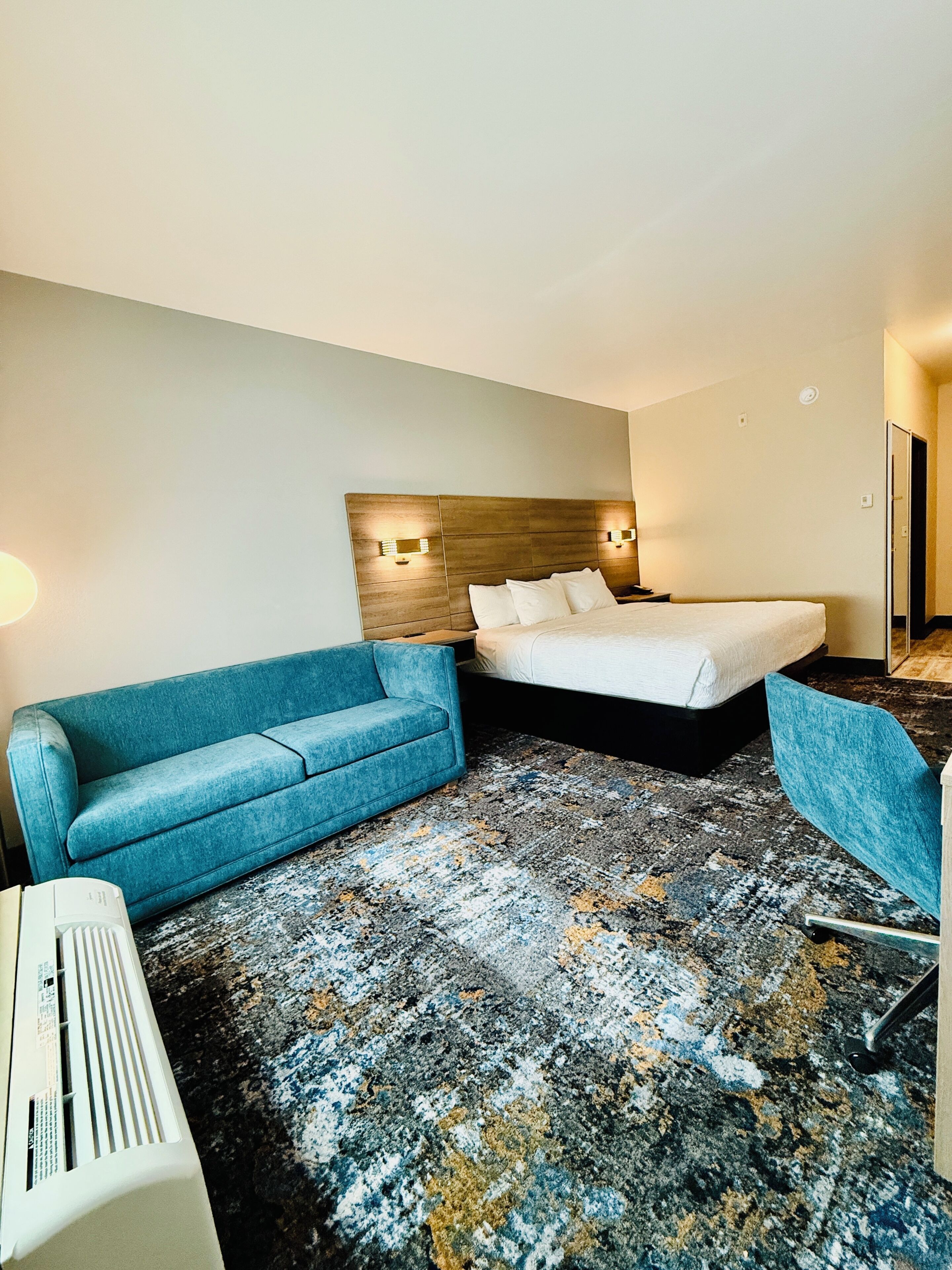 A hotel room with a bed, a blue sofa, a chair, and a heater.
