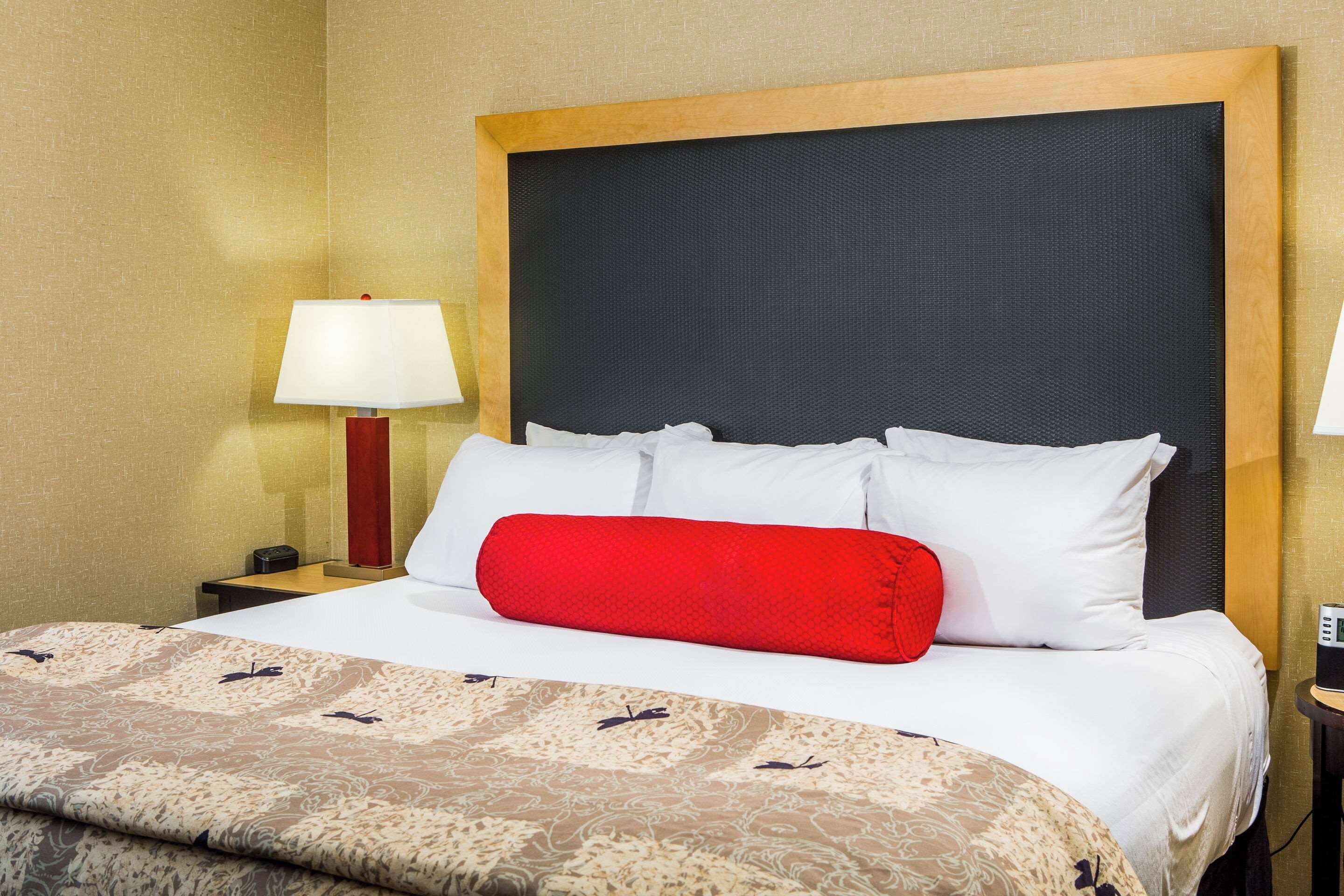 A hotel room with a large bed, a wooden headboard, a red pillow, white bedding, and a lamp on a bedside table.