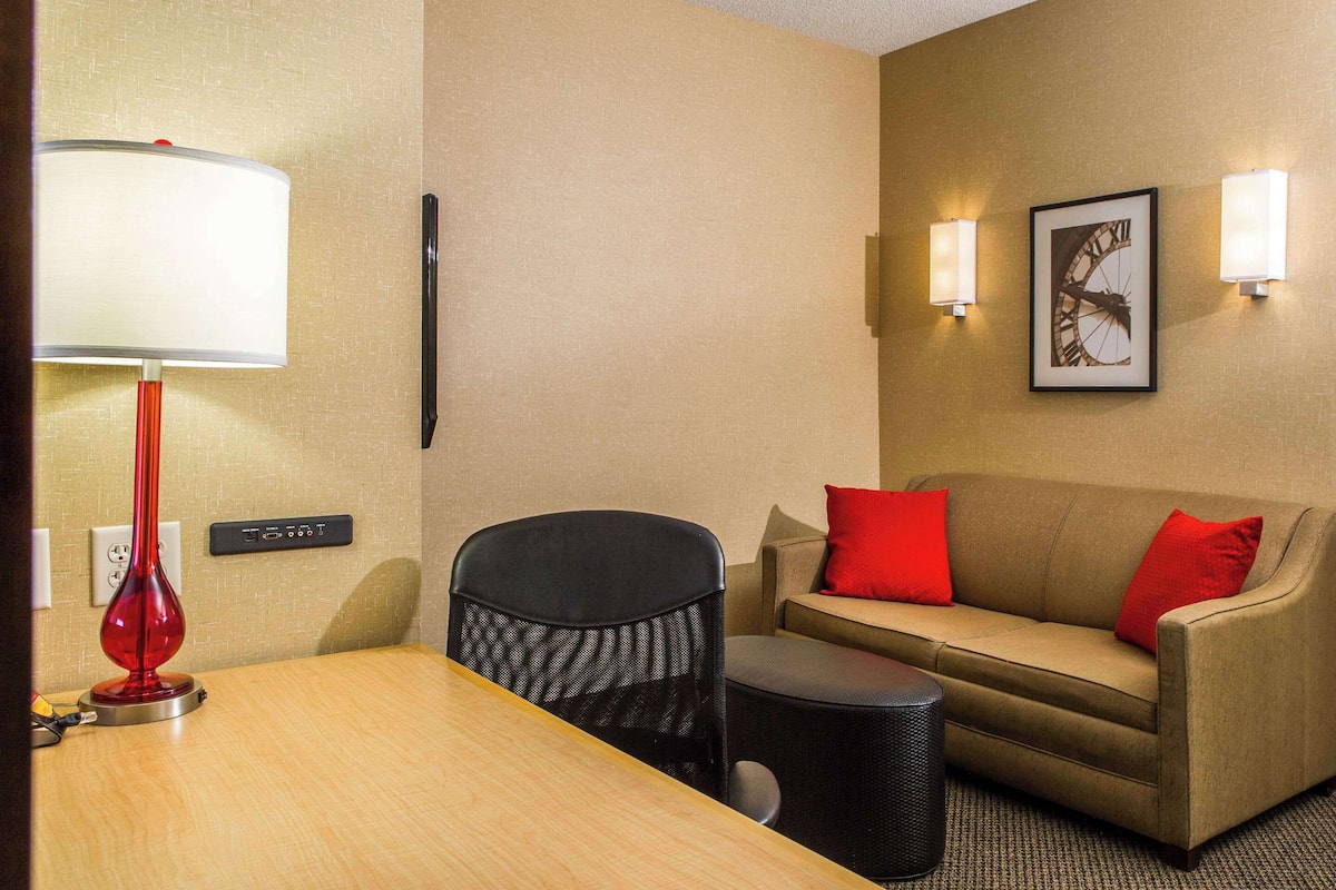 Habitación de hotel con una mesa de madera, una lámpara roja, una silla negra, un sofá color beige con cojines rojos y dos apliques de pared.