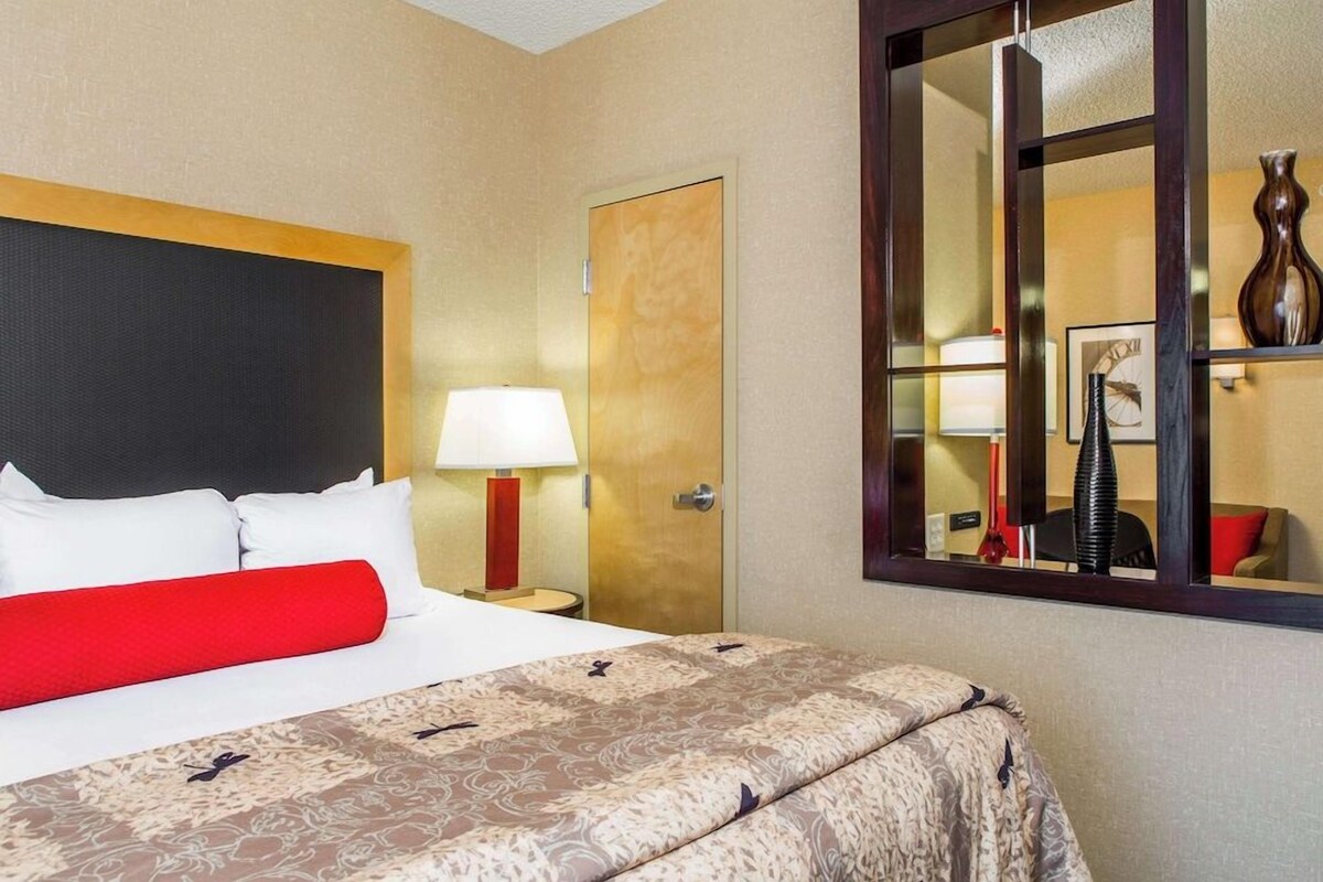A hotel room with a bed, a nightstand with a lamp, a door, and a mirror reflecting part of the room.