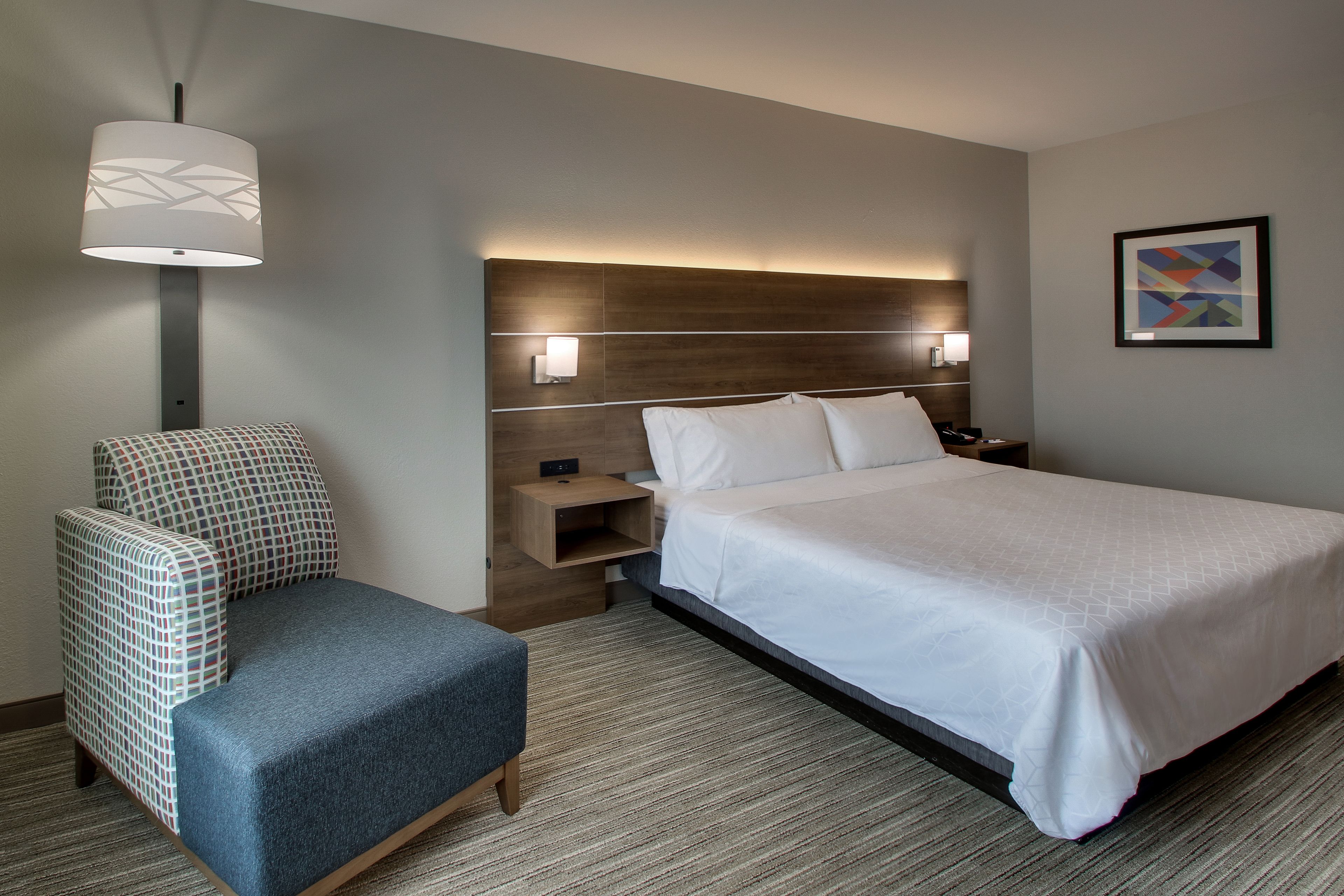 Una habitación de hotel con cama, una silla, una mesita de noche, una lámpara y luz en la pared.