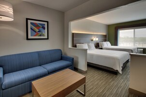 Quarto de hotel com um sofá azul, uma mesa de madeira, uma cama com lençóis brancos, uma cabeceira com luzes fixadas na parede e um quadro abstrato emoldurado.