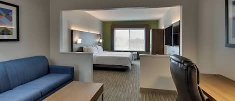 A hotel room with a blue sofa, a wooden coffee table, a black leather chair, a bed with white bedding, a flat-screen TV mounted on the wall, and a large window.