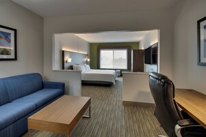 Habitación de hotel con un sofá azul, una mesa de centro de madera, un sillón de cuero negro, una cama con ropa de cama blanca, un televisor de pantalla plana montado en la pared y un ventanal grande.