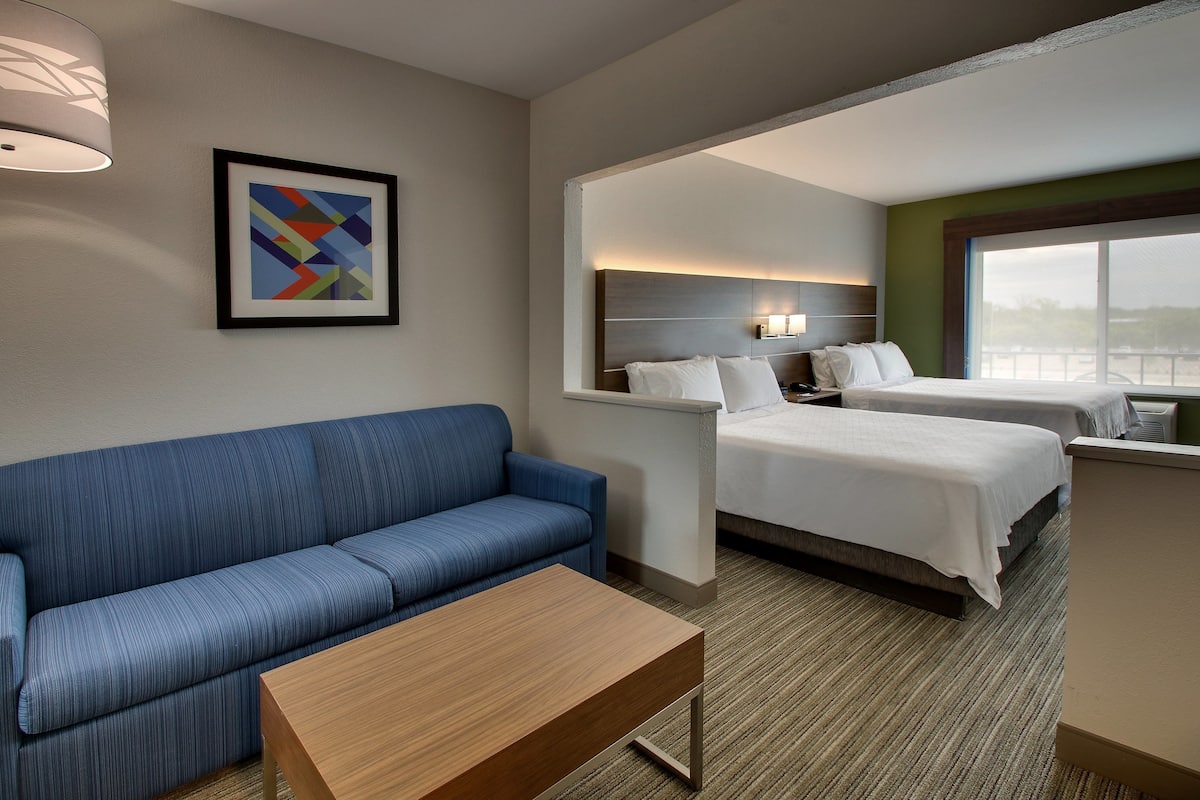 A hotel room with a blue sofa, a wooden table, two beds, a nightstand, and a framed abstract painting.