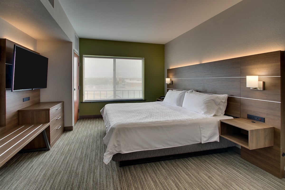 A modern hotel room with a large bed, a flat-screen TV, a desk with drawers, and a window with a view.