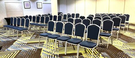 A conference room with rows of chairs and tables, patterned carpet, and a plain wall with framed artwork.