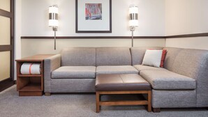 Een moderne hotelkamer met een grijze hoekbank, een houten bankje en een klein tafeltje met een handdoek. Twee wandlampen en een lijst met een afbeelding zijn zichtbaar.