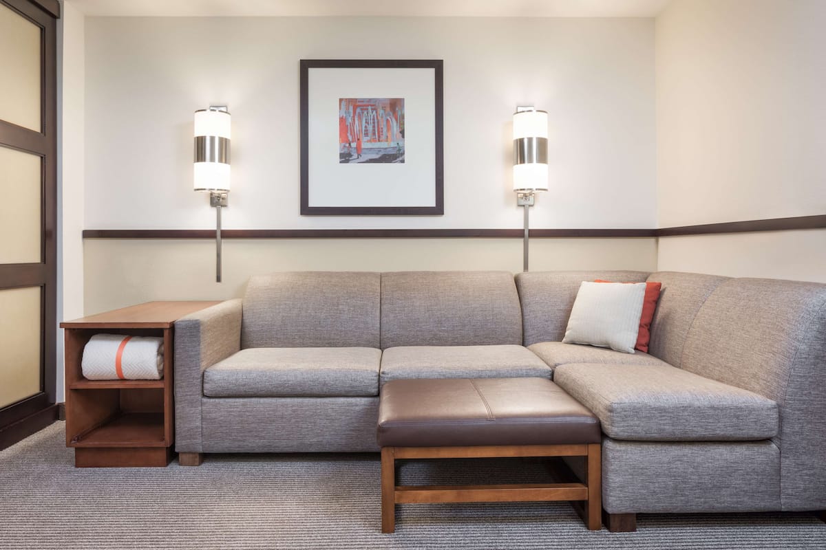 Ein modernes Hotelzimmer mit einem grauen Ecksofa, einer Holzbank und einem kleinen Tisch mit einem Handtuch. Zwei Wandlampen und ein gerahmtes Bild sind sichtbar.