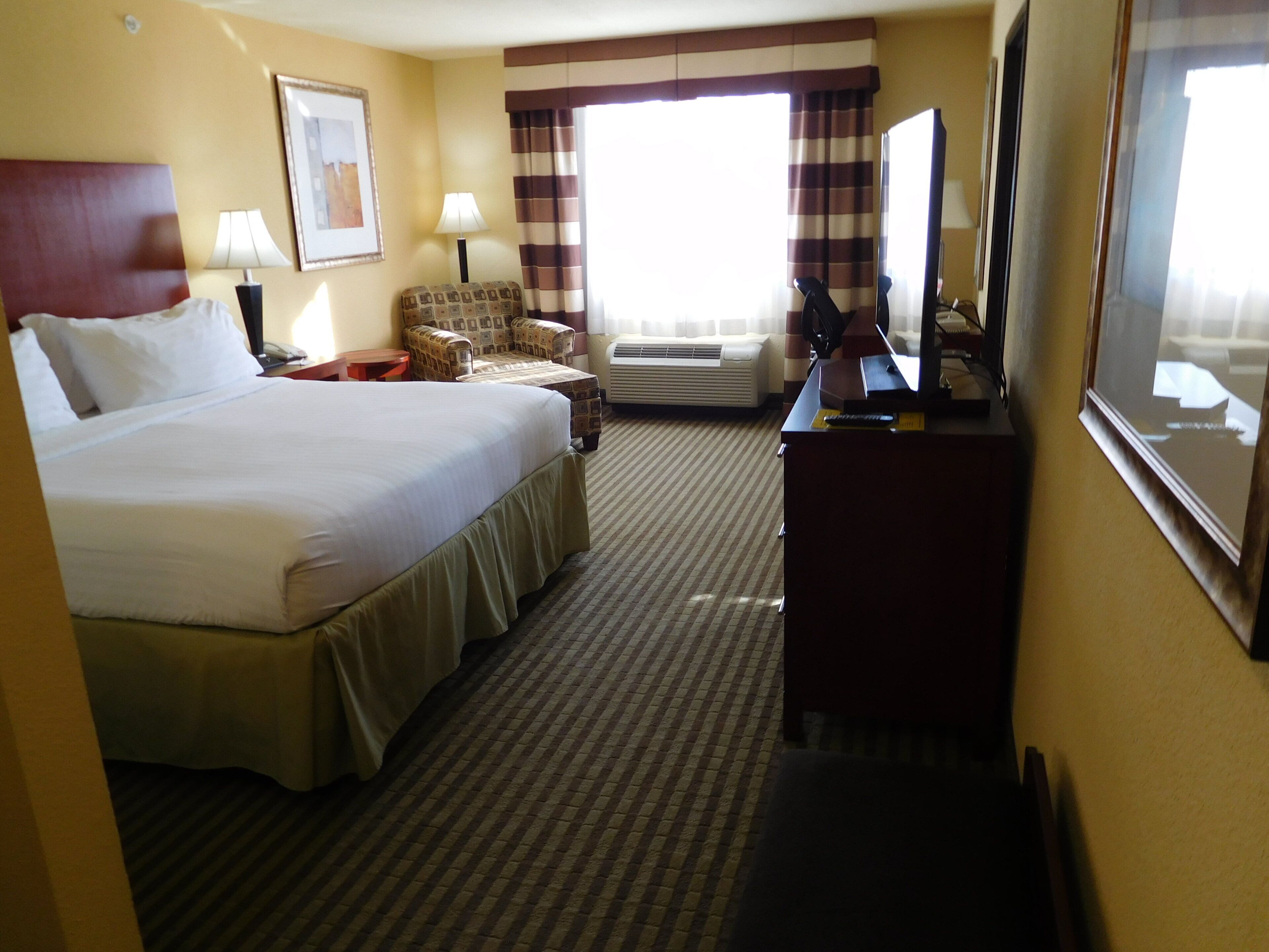 A hotel room with a large bed, a desk, a chair, a television, and an air conditioning unit.
