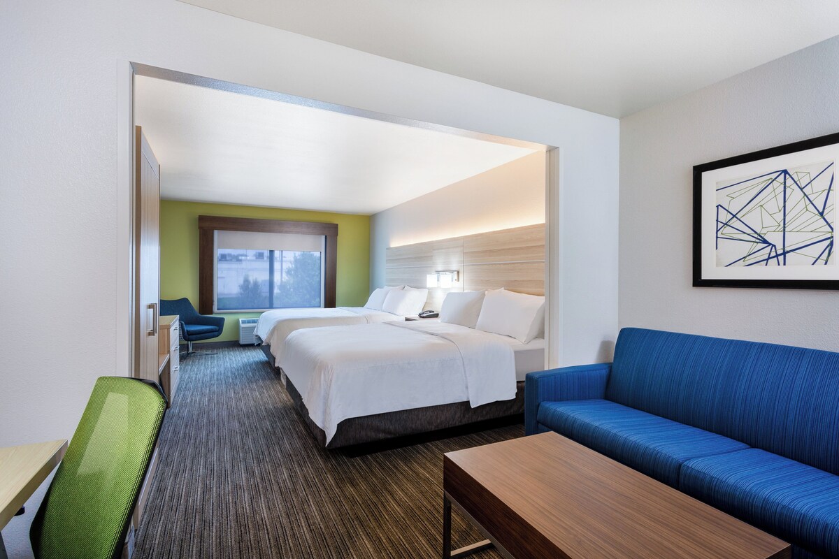 Habitación de hotel con dos camas, un sofá azul, una mesa de centro de madera y un cuadro abstracto enmarcado en la pared.