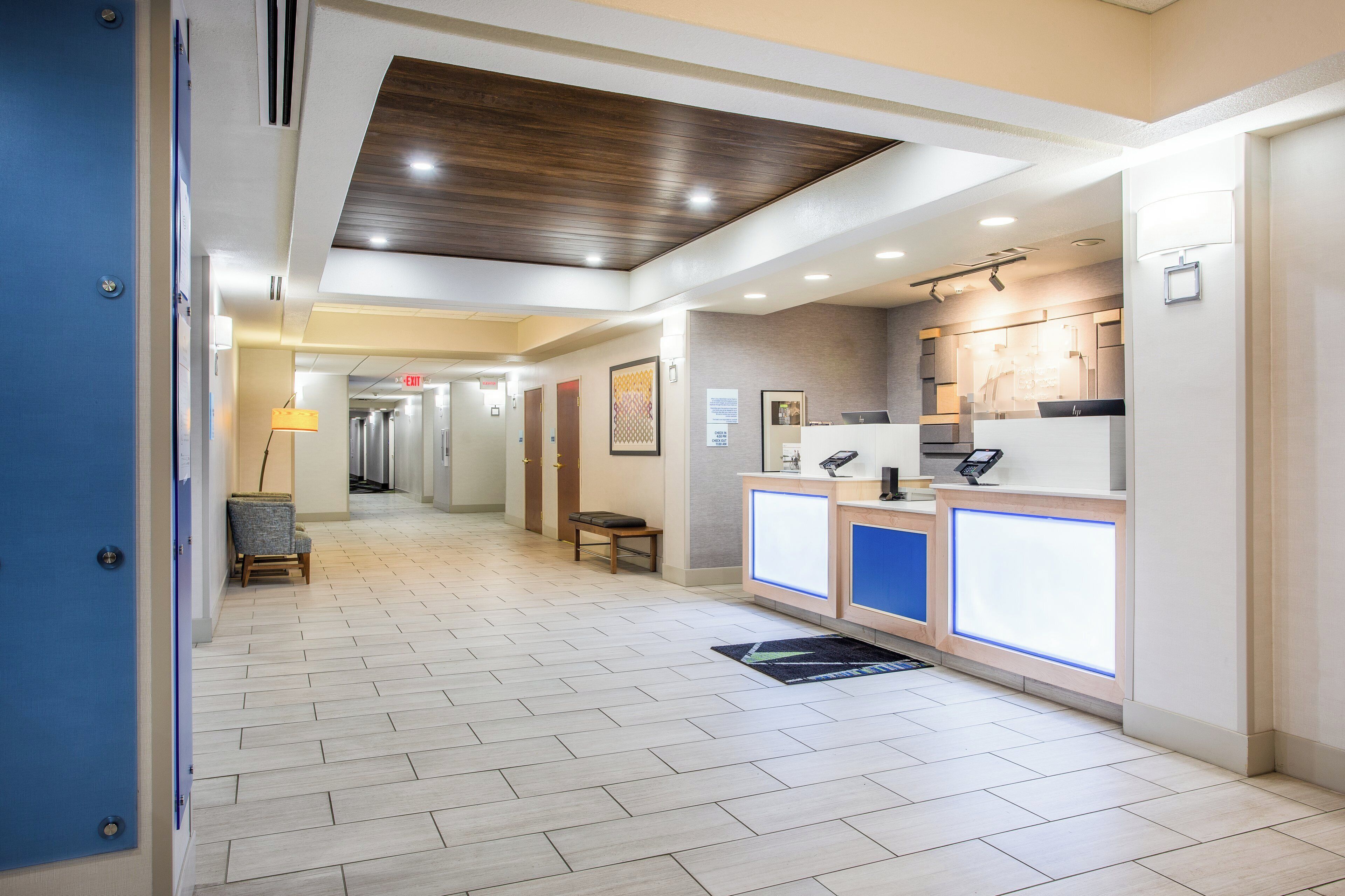 A hotel reception area with a check-in counter, a waiting area with a bench, and a hallway leading to elevators.