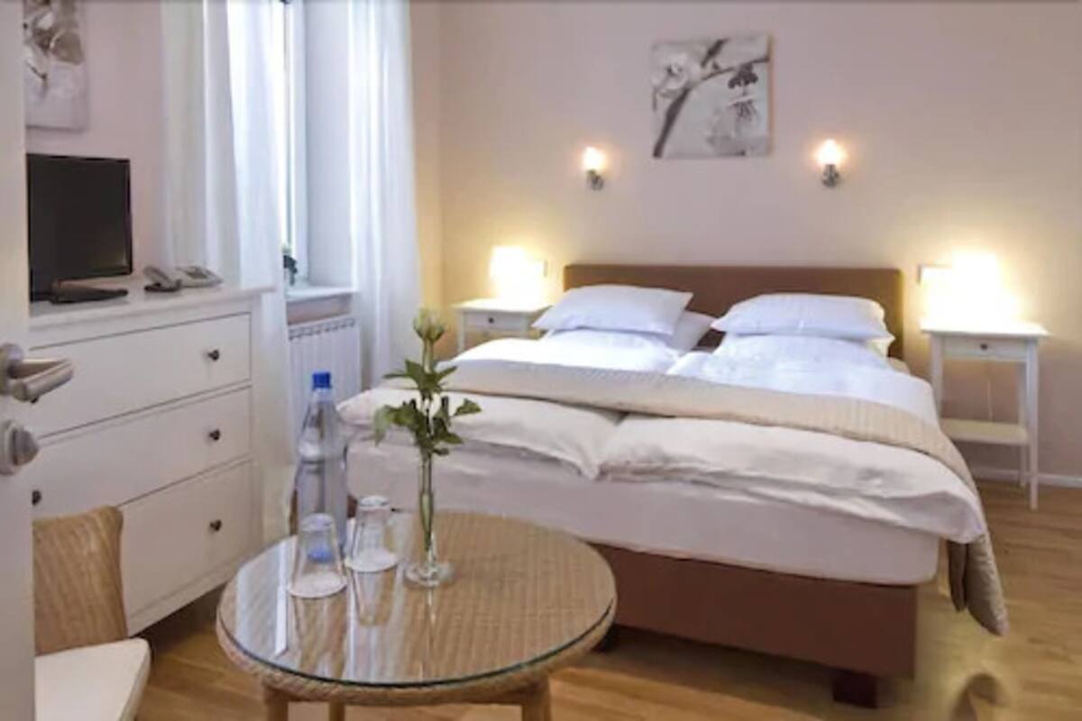 Ein Hotelzimmer mit einem Bett, Nachttischen, einem Glastisch mit einer Vase, einem Sessel und einem Fernseher auf einer Kommode.