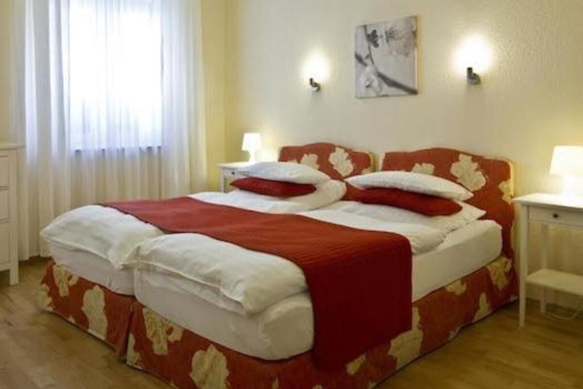 A double bed with a red and white bedspread, two bedside tables with lamps, a window with white curtains, and a wall-mounted artwork.