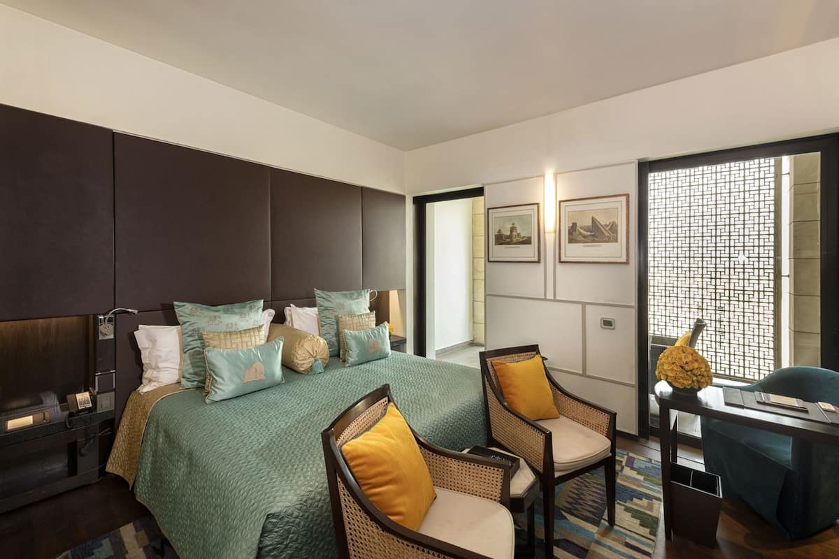 A modern hotel room with a large bed, two chairs, a desk, and a view through a window with a geometric pattern.