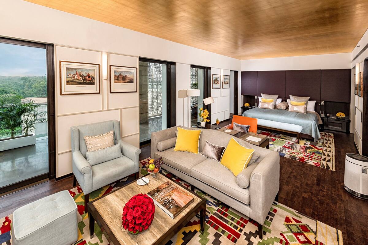 A modern hotel room with a large bed, a sofa, a coffee table with a book and flowers, and a view of greenery through the window.