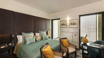 A modern hotel room with a large bed, two chairs, a desk, and a view through a window with a geometric pattern.