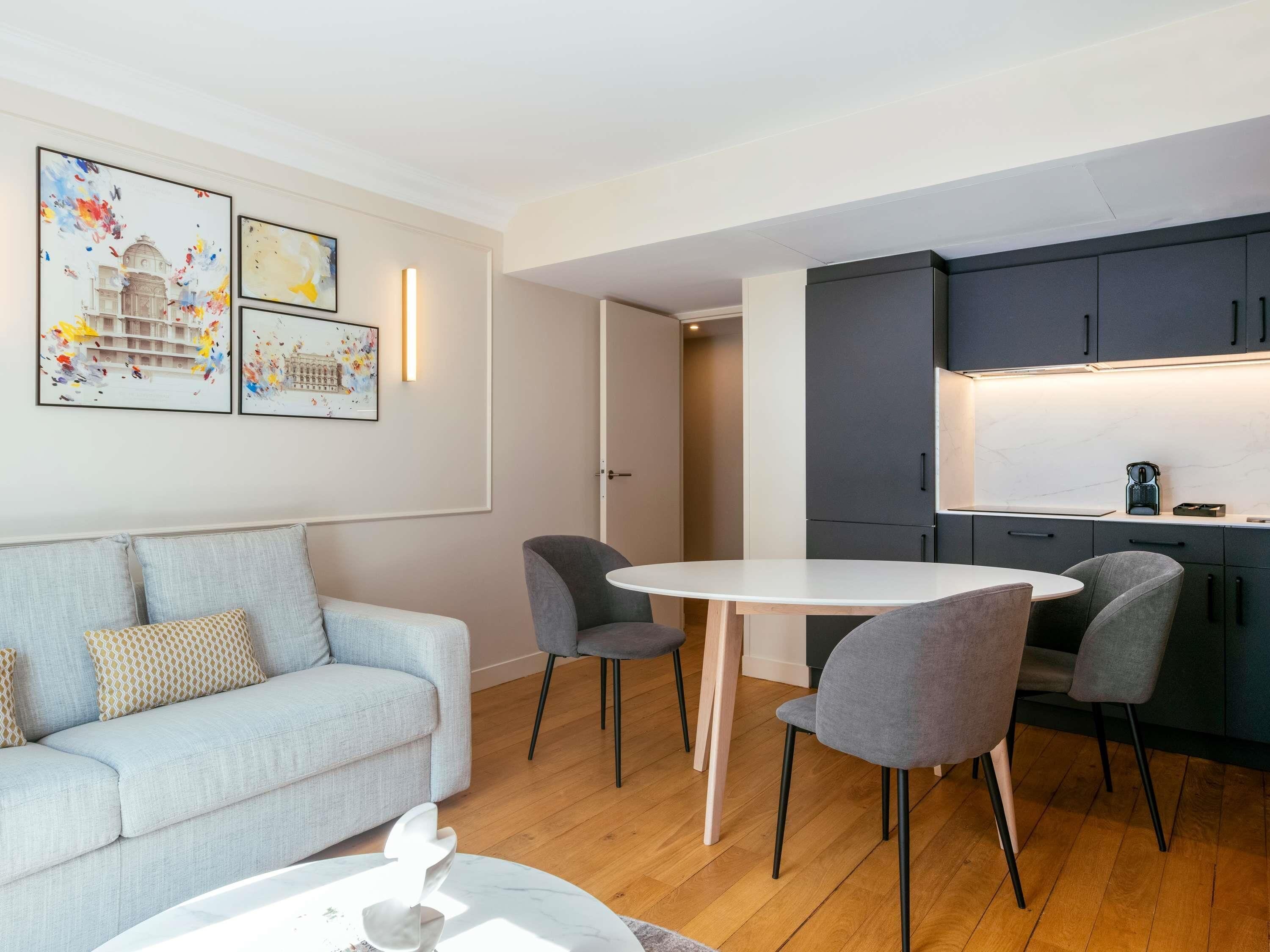 A modern living room with a sofa, a round dining table, and chairs, a kitchen area with dark cabinets and a countertop, and two framed artworks on the wall.