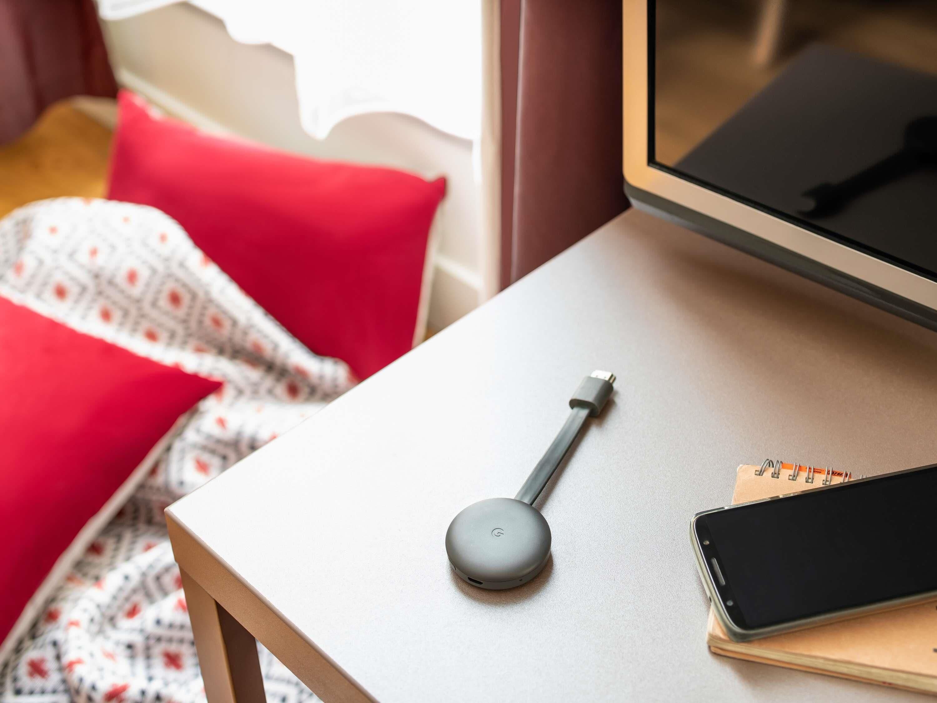 A bed with a red pillow, a blanket, a bedside table with a smartphone and a charging device.