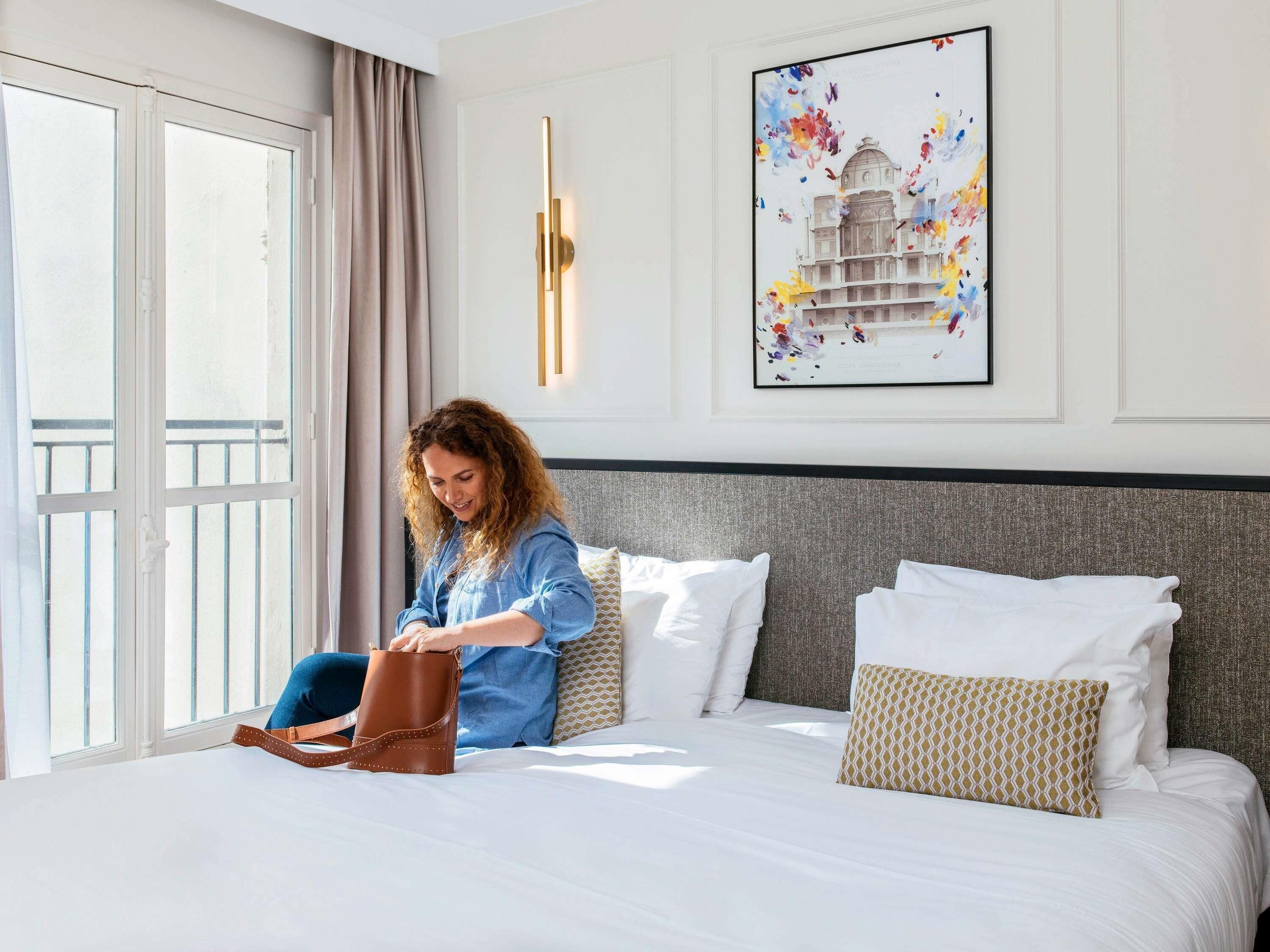 A hotel room with a bed, a window with curtains, a wall-mounted light, and a framed picture.