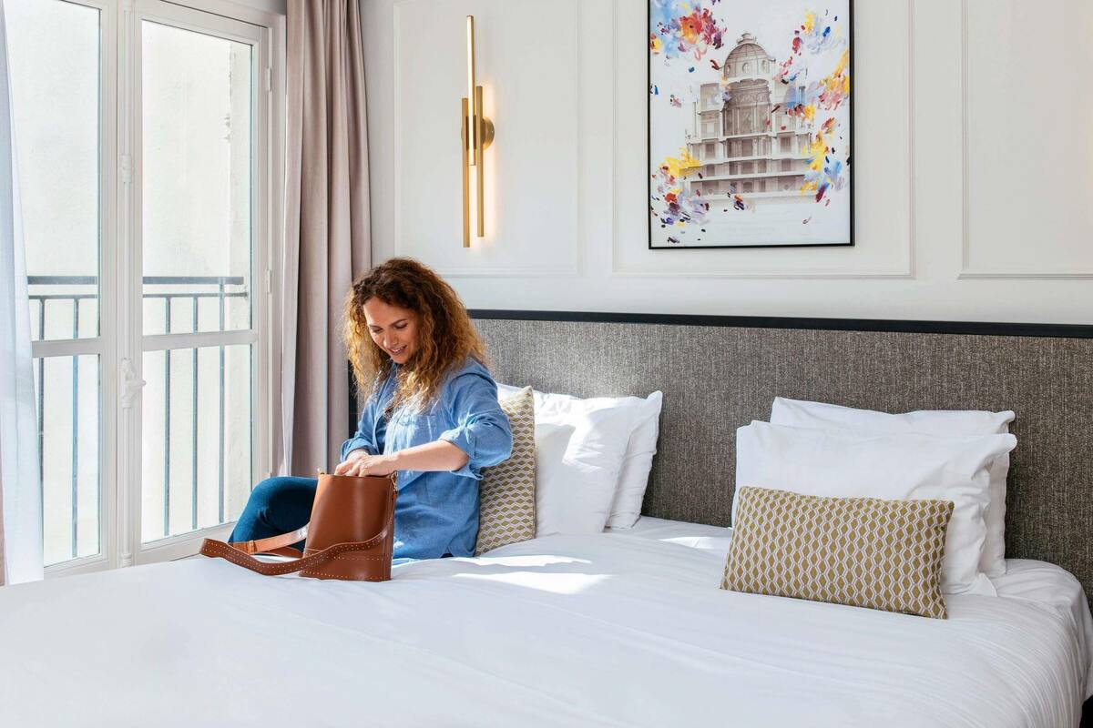 Una habitación de hotel con una cama, un ventanal con cortinas, una lámpara de pared y un cuadro enmarcado.