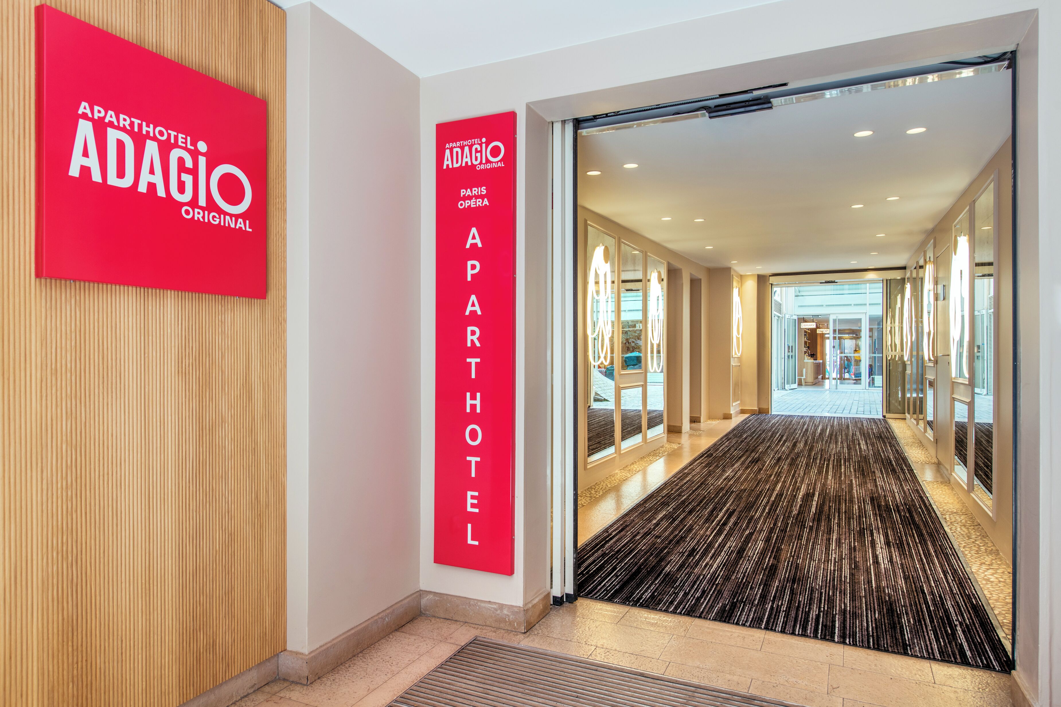 L’entrée d’un hôtel, avec une enseigne rouge indiquant « Adagio Paris Op éra », un intérieur moderne et un couloir recouvert de moquette.