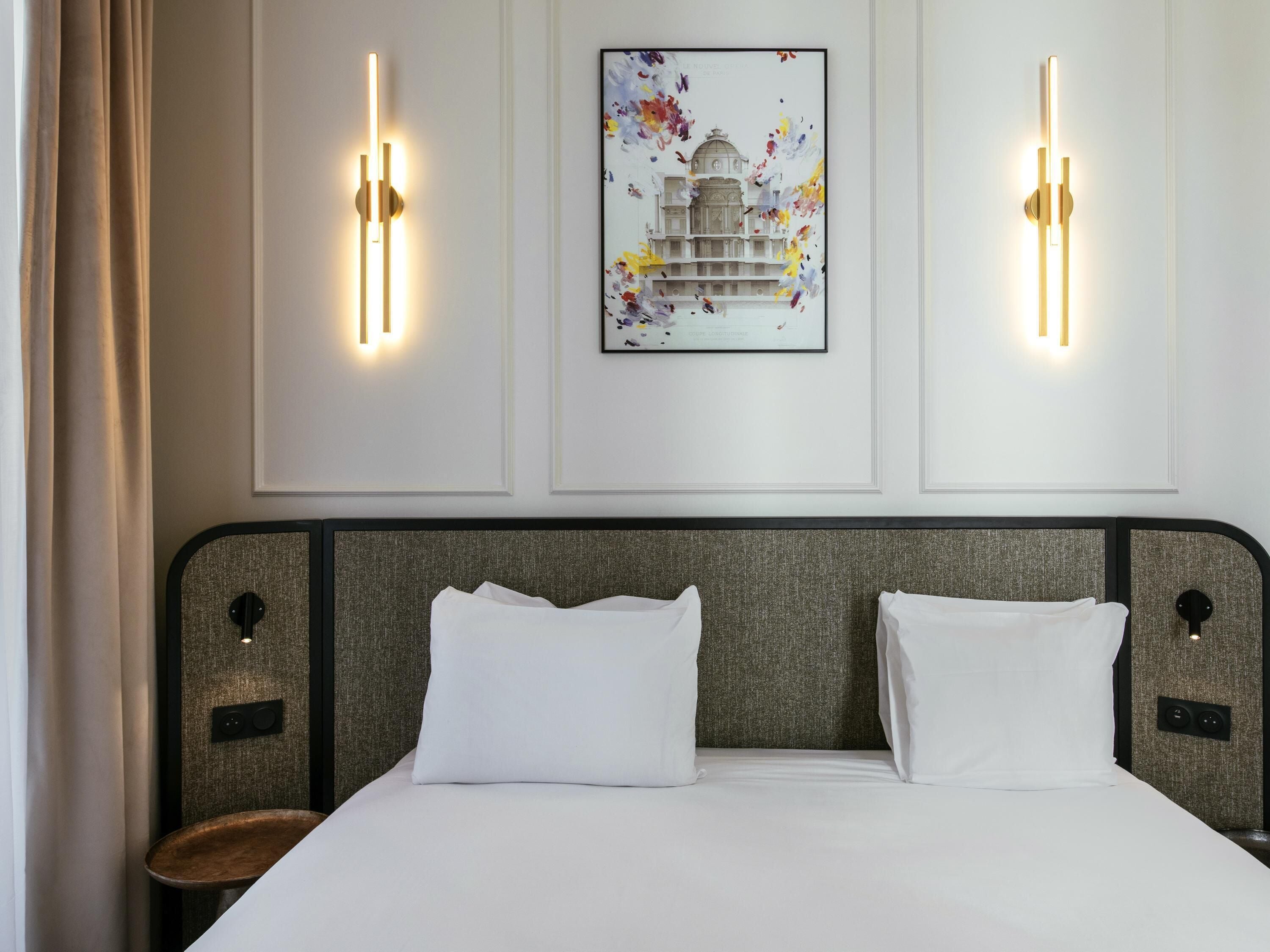 Hotellihuone, jossa on suuri sänky, kaksi seinälle asennettua valaisinta ja sängyn yläpuolella oleva kehystetty taulu.