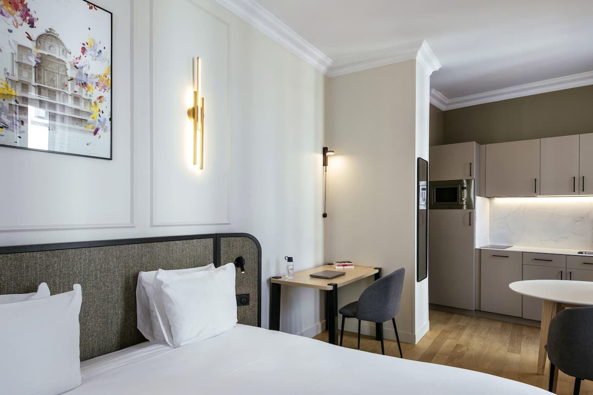 A hotel room with a bed, a desk, a chair, a microwave, and a kitchen area.