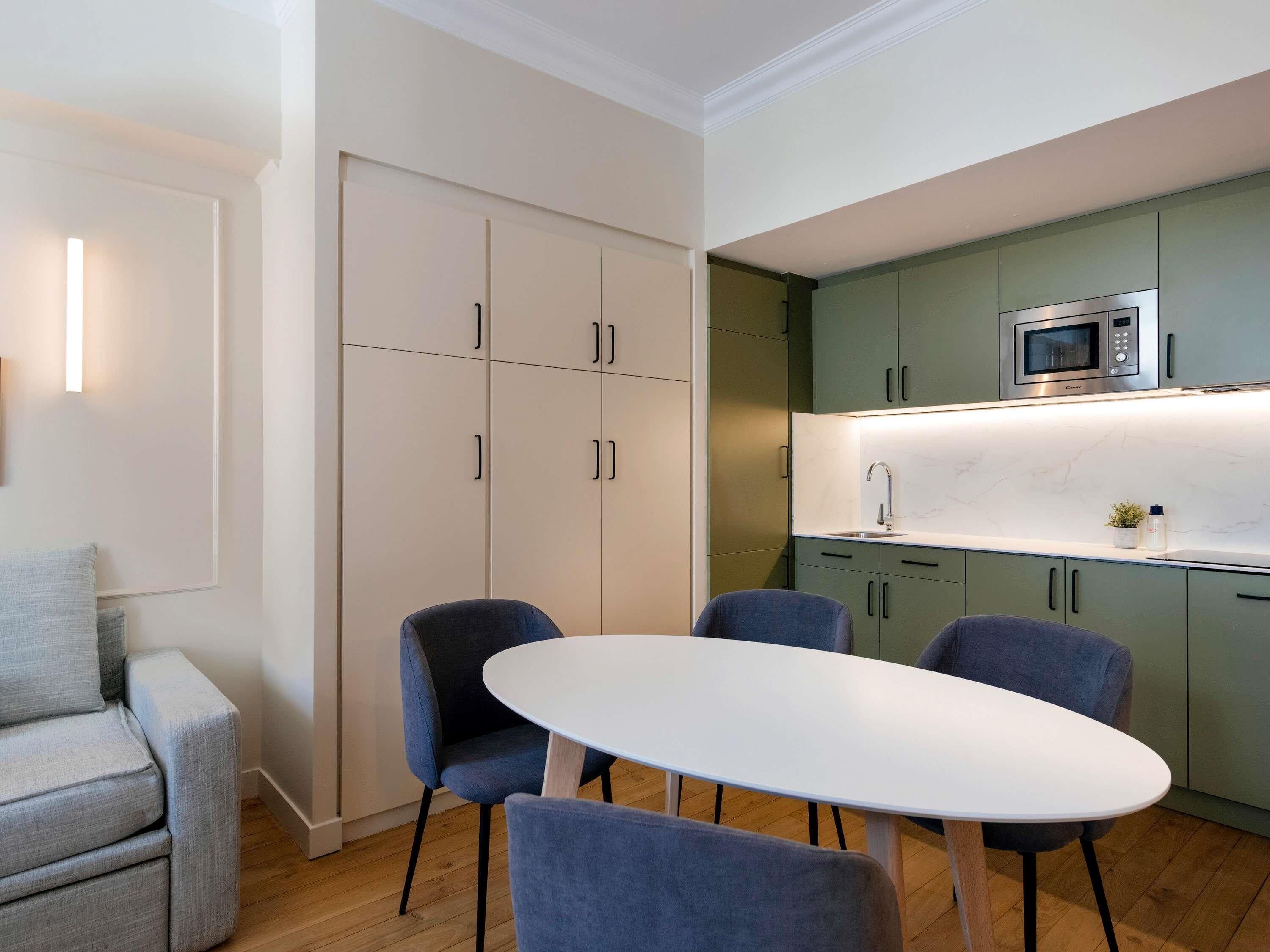 Cocina moderna con encimera blanca, microondas integrado y un lavabo. Hay dos sillas azules alrededor de una mesa blanca.