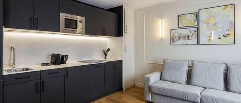 Une cuisine moderne avec des meubles de couleur sombre, un four à micro-ondes encastré et une machine à café. Le salon est équipé d’un canapé gris et d’une table basse ronde.