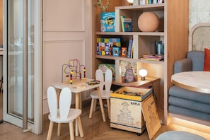 Ein Spielbereich für Kinder mit einem Holztisch, Stühlen, einem Bücherregal mit Büchern und Spielzeug und einer kleinen Lampe.