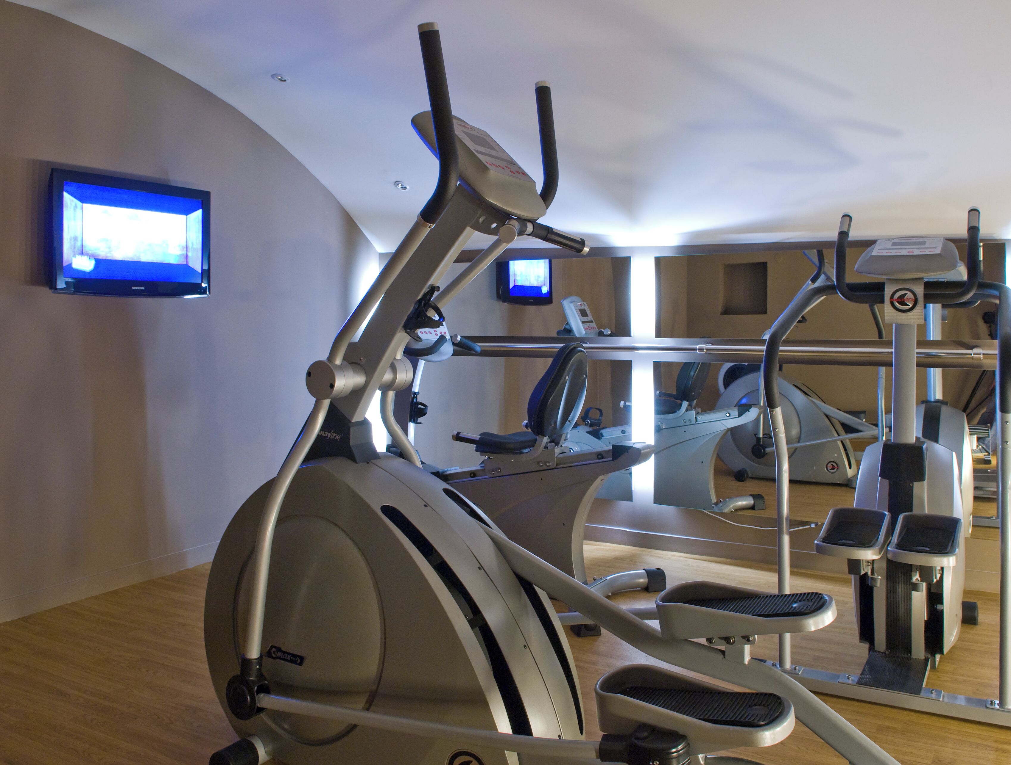 A gym with cardio equipment, a TV mounted on the wall, and a wooden floor.
