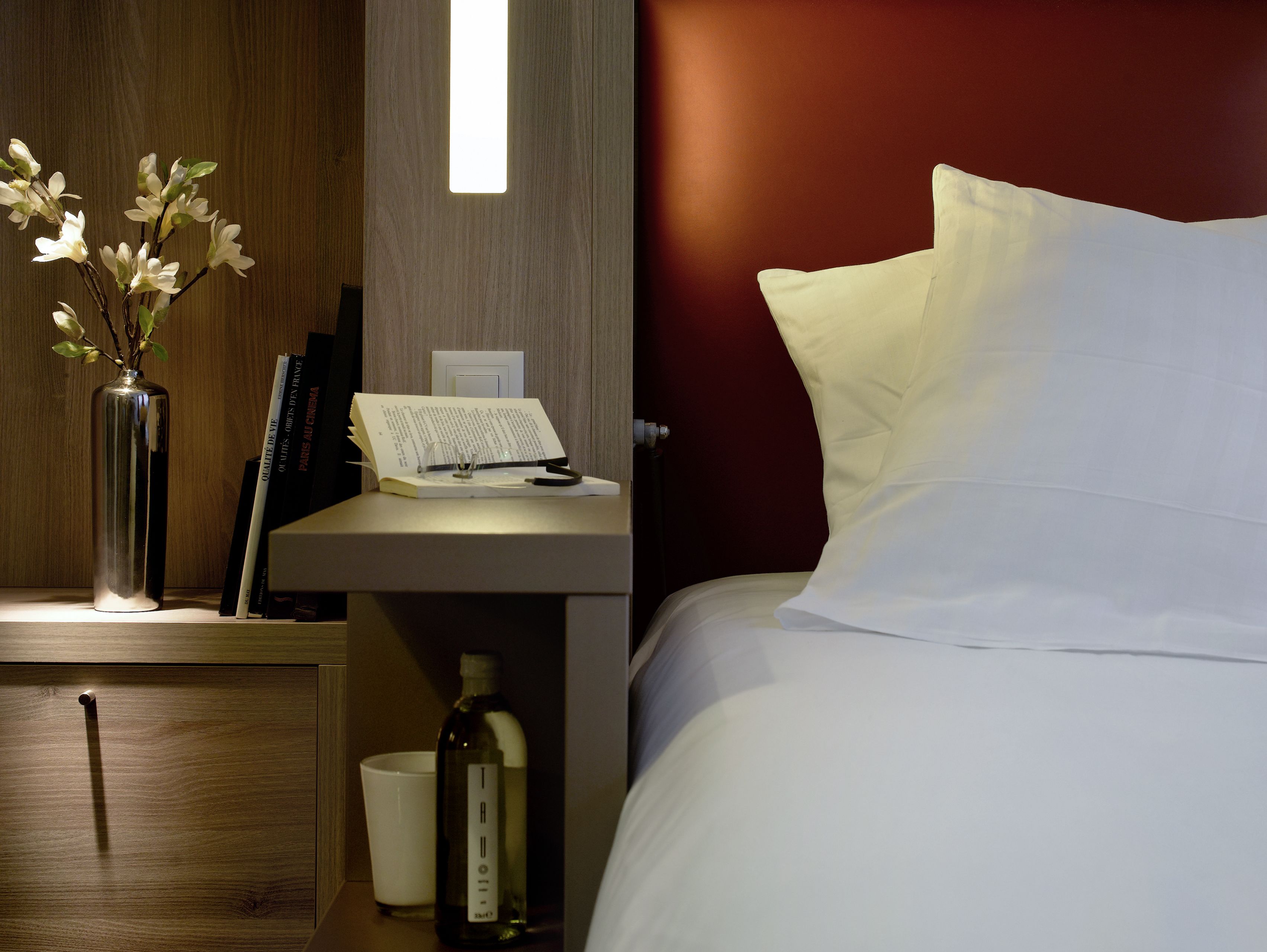 Habitación de hotel con mesita de noche de madera, un florero con flores, un libro, una botella y una cama con ropa de cama blanca.