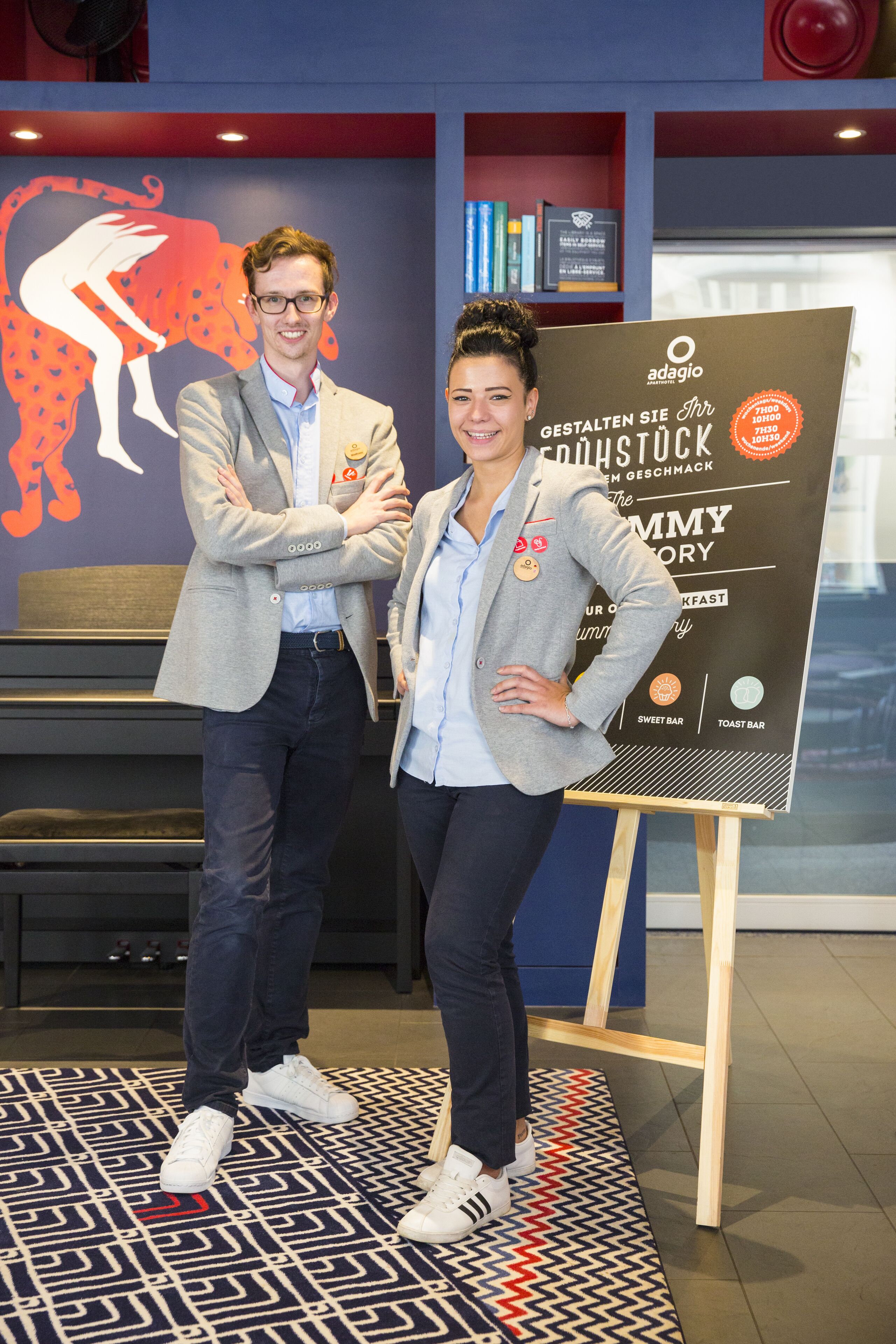 Two people standing in front of a sign with text and logos, in a room with a patterned floor and a bench in the background.