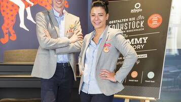 Two people standing in front of a sign with text and logos, in a room with a patterned floor and a bench in the background.