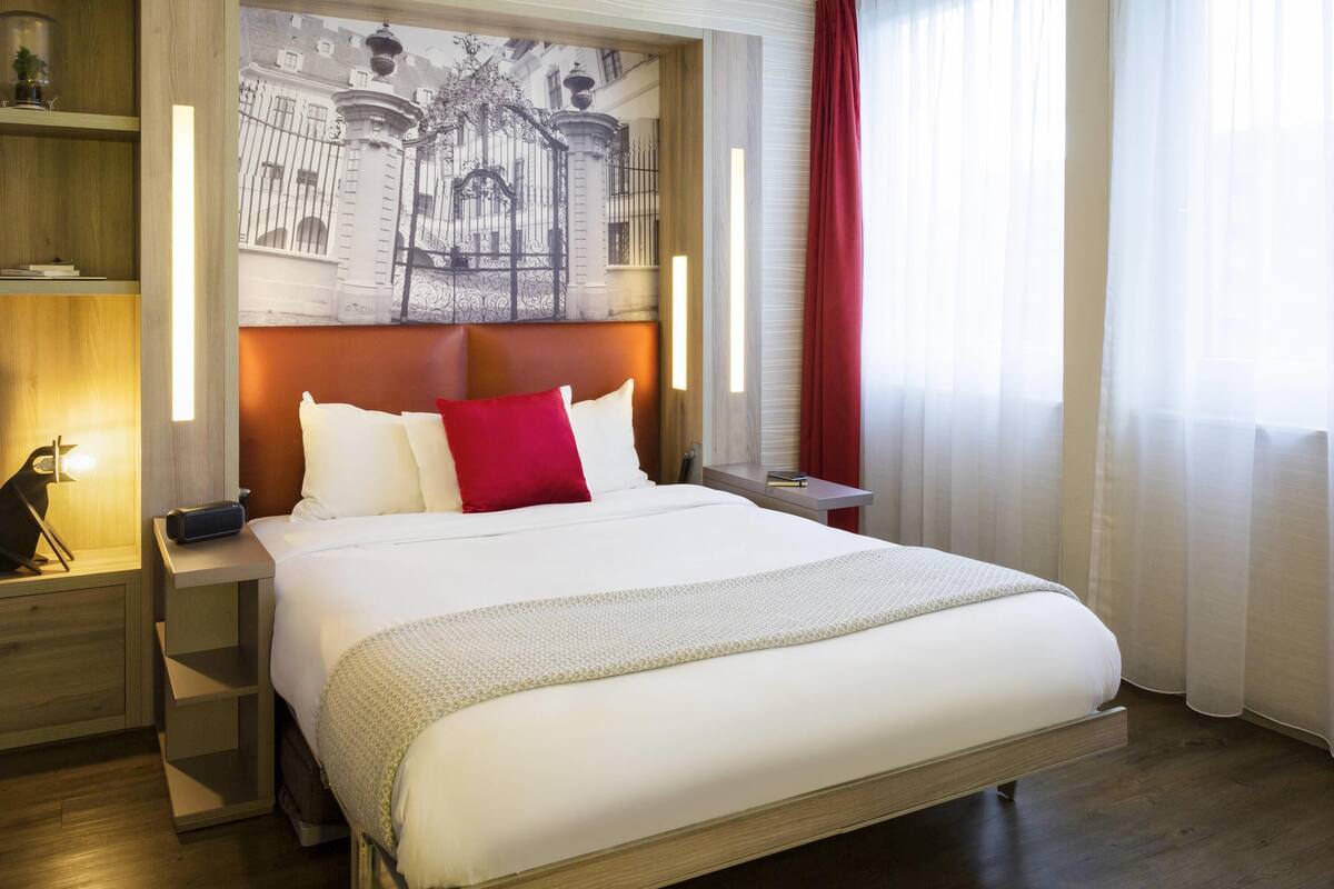 Habitación de hotel con una cama grande, una mesita de noche con lámpara, una estantería y un amplio ventanal con cortinas.
