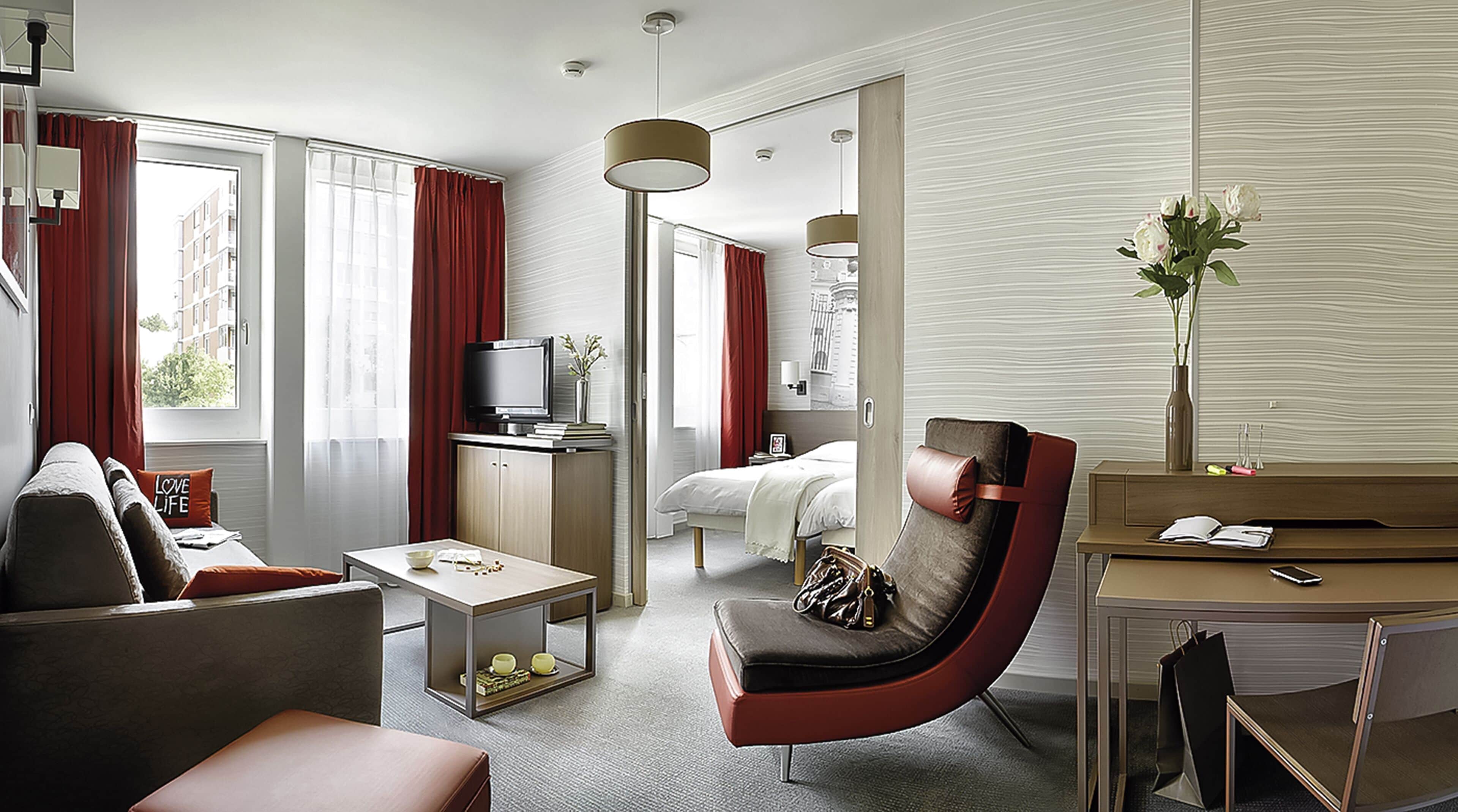 A modern hotel room with a sofa, a chair, a desk, a bed, a TV, and a large window with curtains.