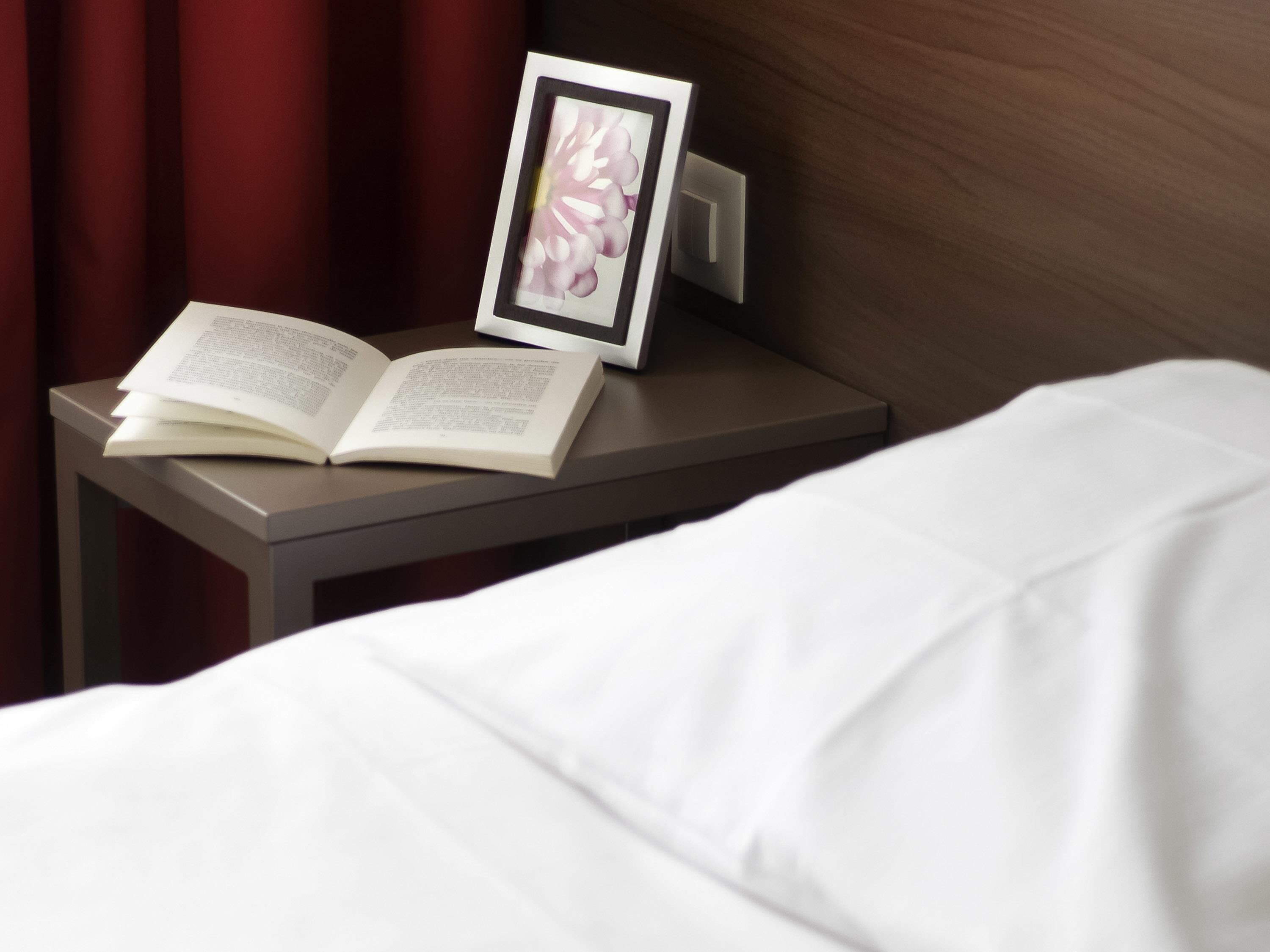 Een net opgemaakt bed met een witte deken, een nachtkastje met een boek en een ingelijste foto, en een rood gordijn op de achtergrond.