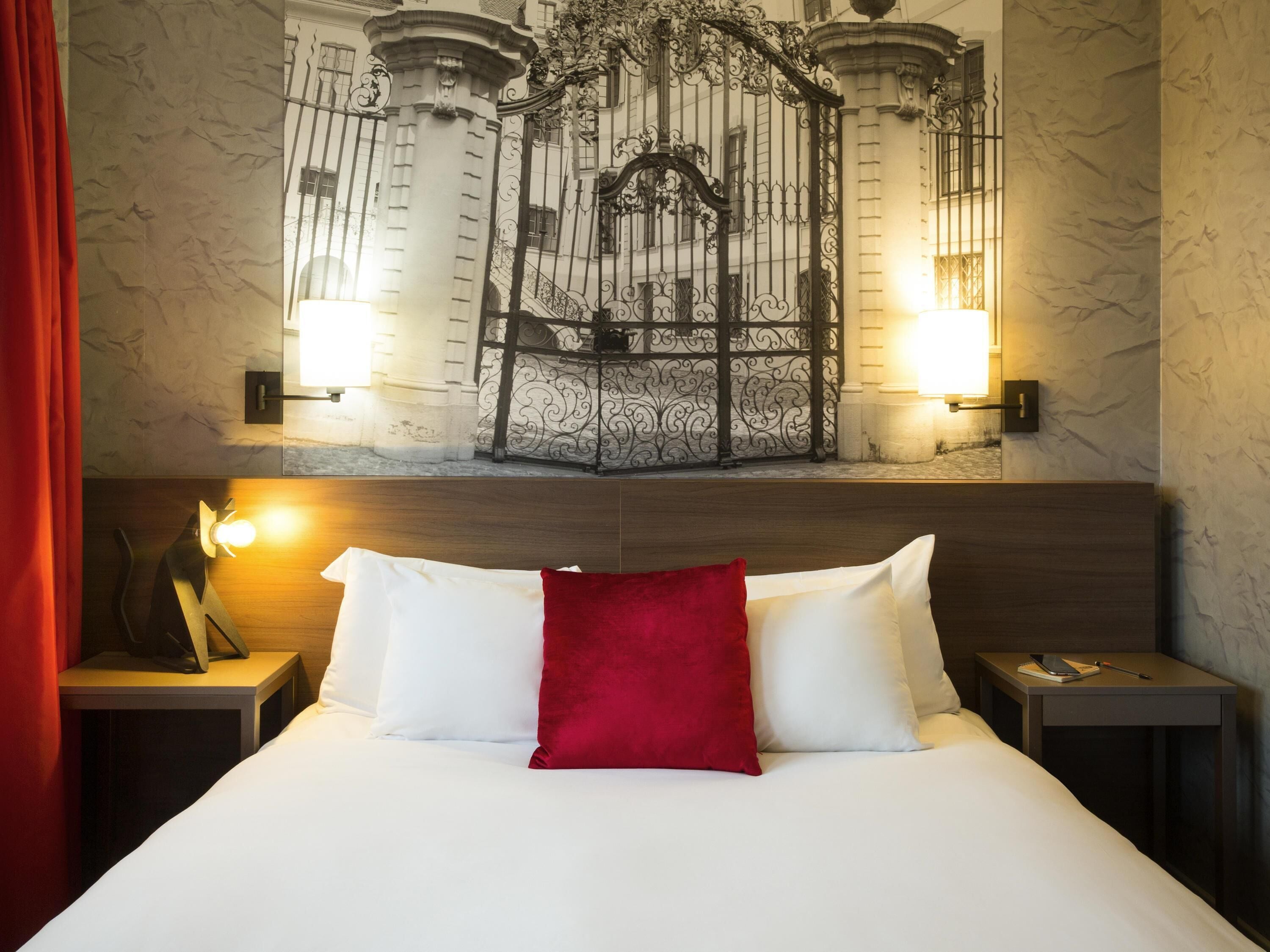 A hotel room with a bed, two bedside tables, a red pillow, and a wall-mounted artwork.