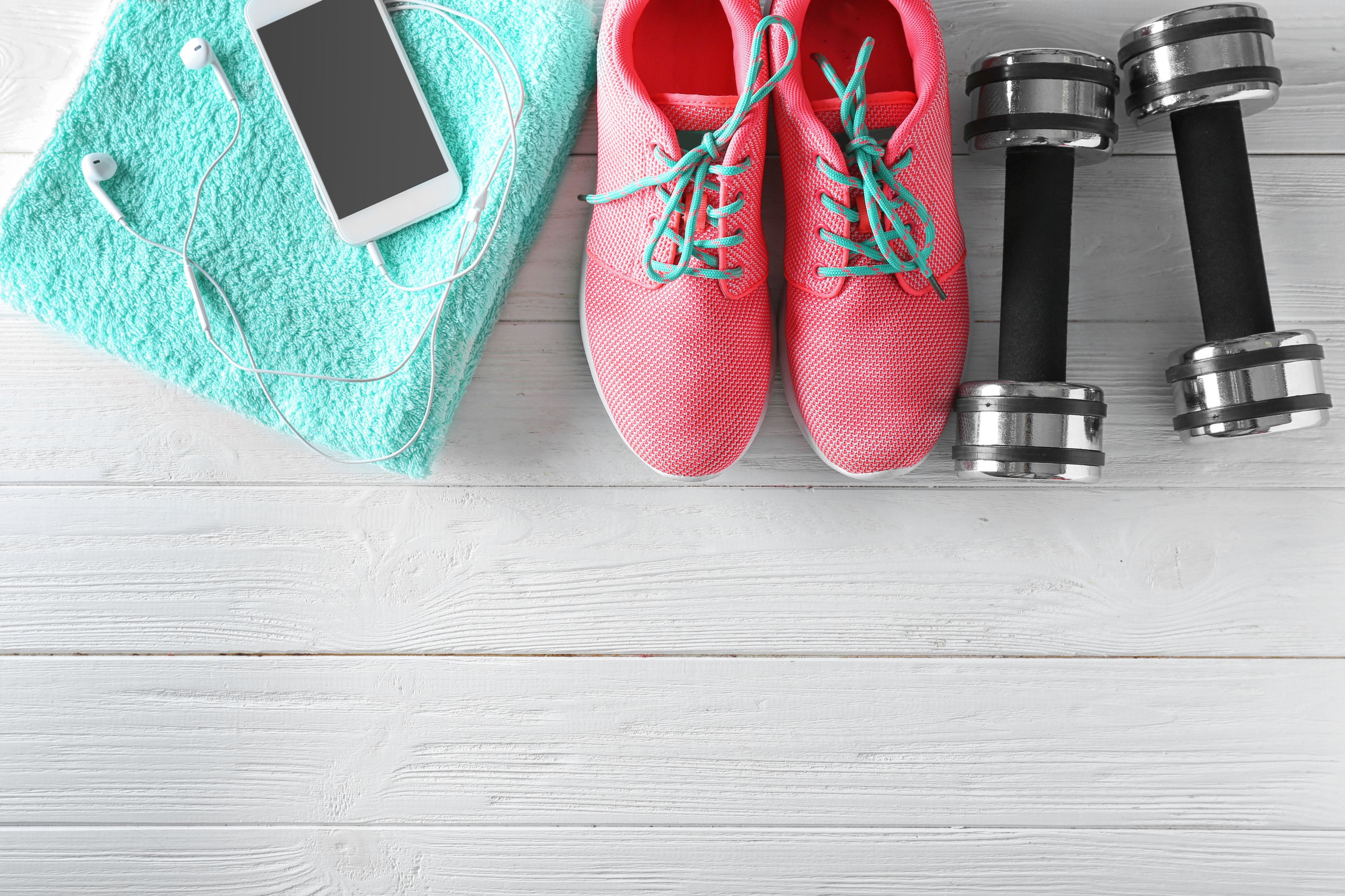 A pair of red sneakers, black dumbbells, a smartphone, and a turquoise towel on a wooden surface.