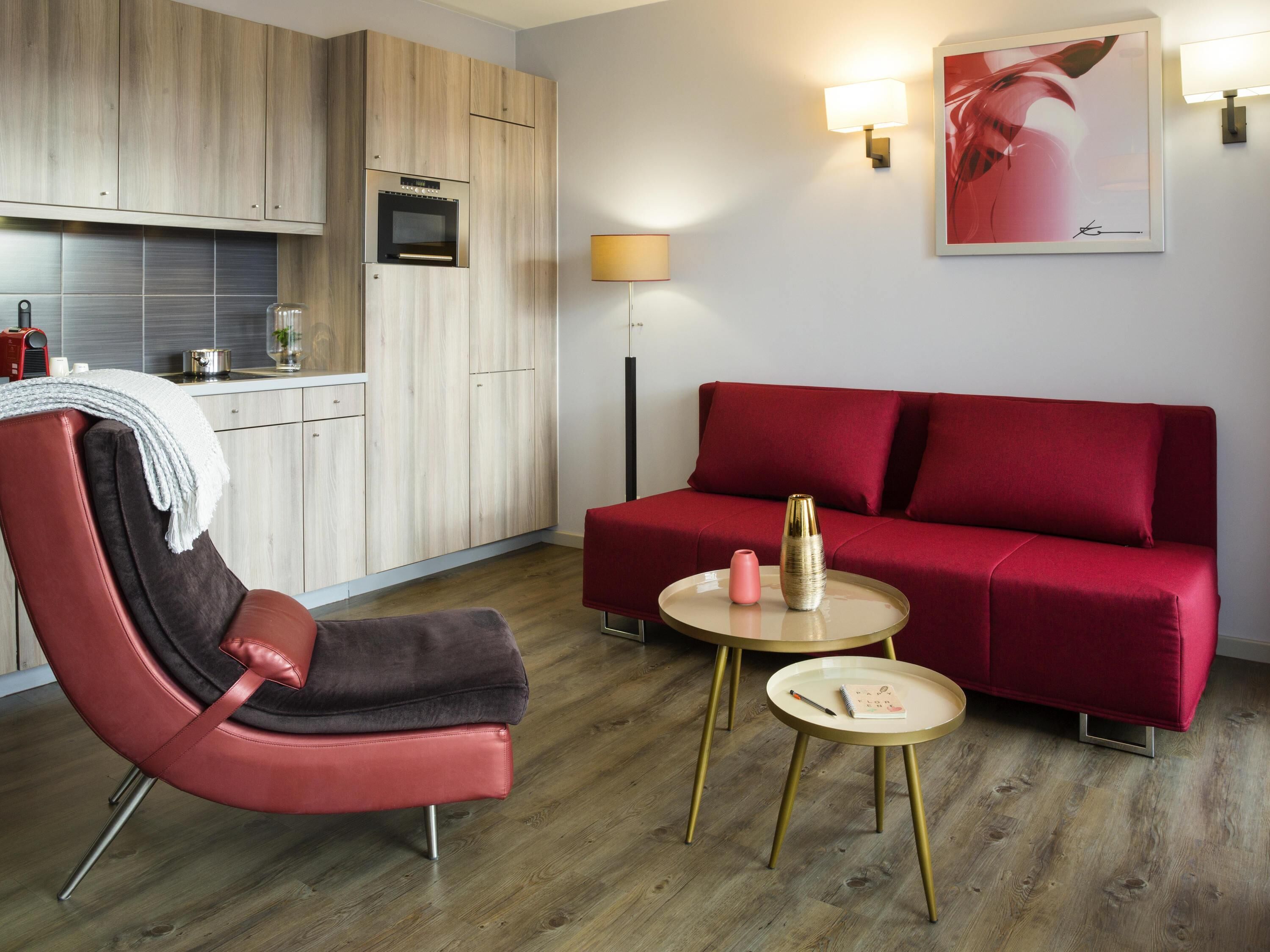 Un espace de vie moderne comprenant un canapé rouge, un fauteuil rouge, une petite table ronde, un coin cuisine avec des meubles en bois et une lampe fixée au mur.