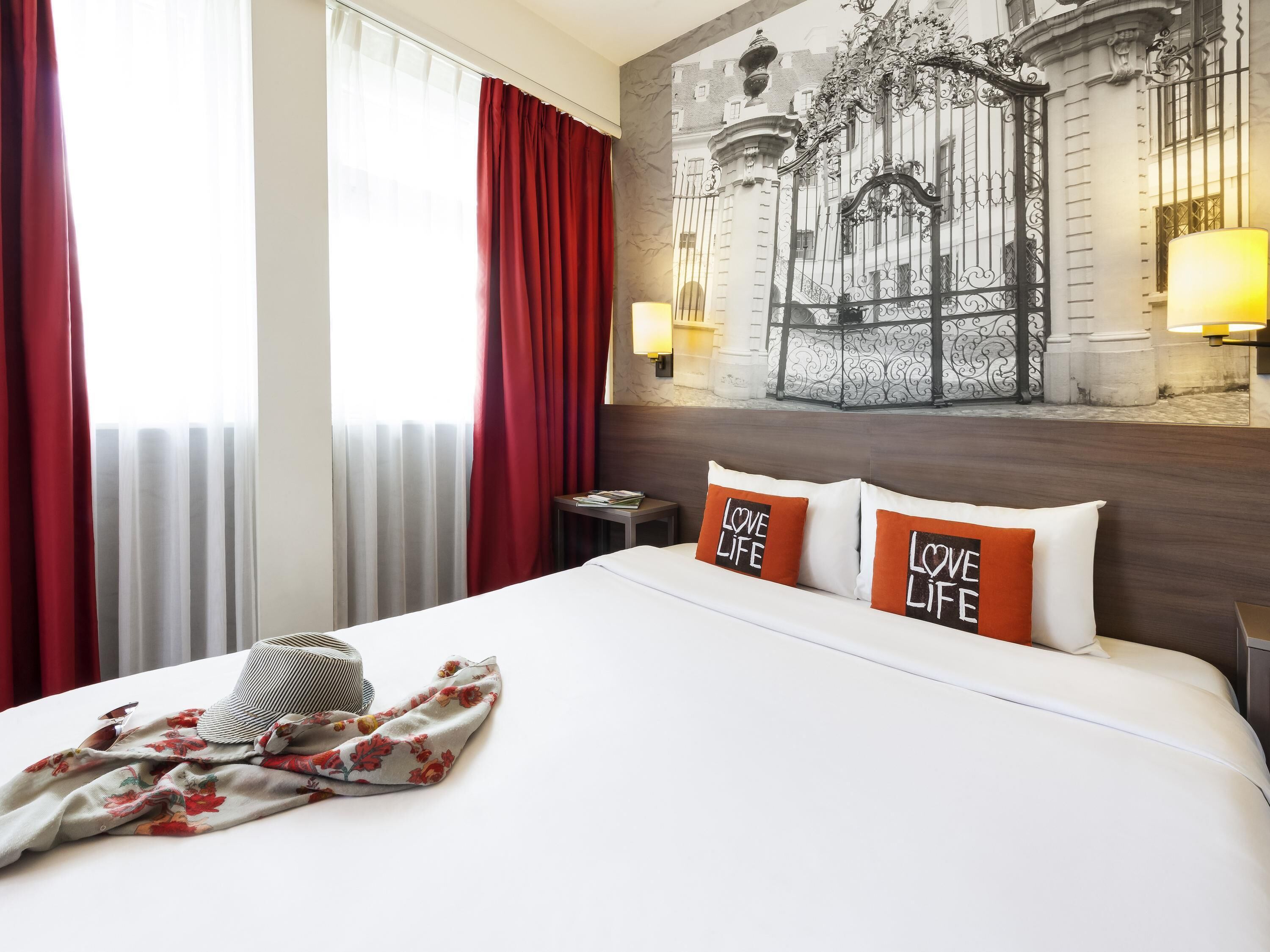 Une chambre d'hôtel avec un lit, des rideaux rouges, une couverture à motifs floraux et deux oreillers ornés de l'inscription « LOVE LIFE ».