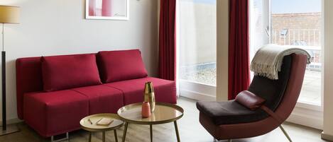 A modern living room with a red sofa, a round coffee table, and a leather armchair. There is a large window with red curtains, a floor lamp, and a wall-mounted light fixture.