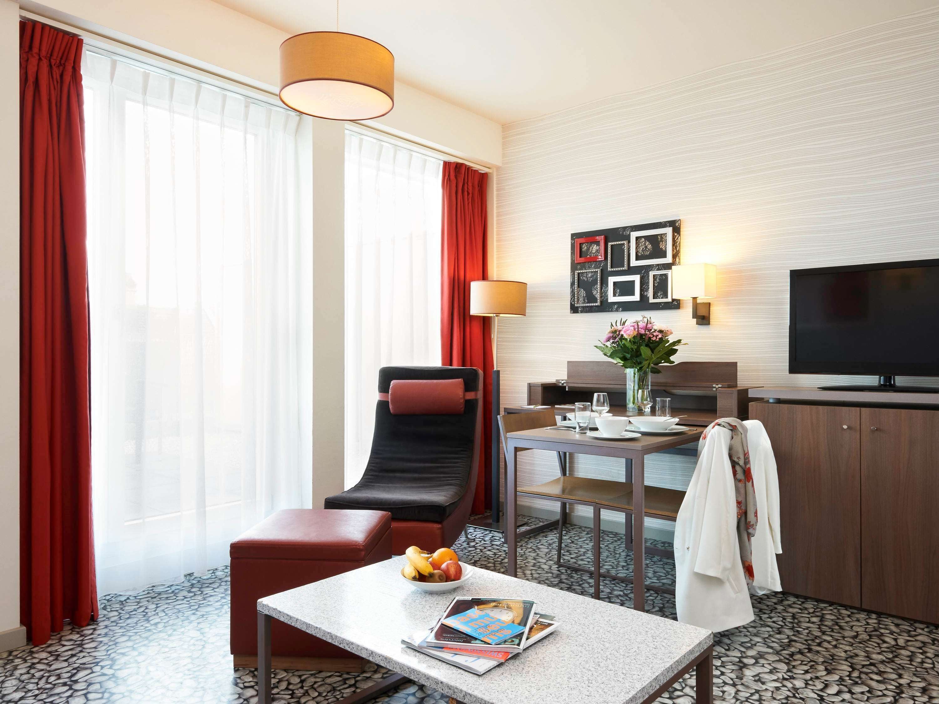 Habitación de hotel con comedor, televisión, sofá y una mesa con un tazón de fruta y revistas.
