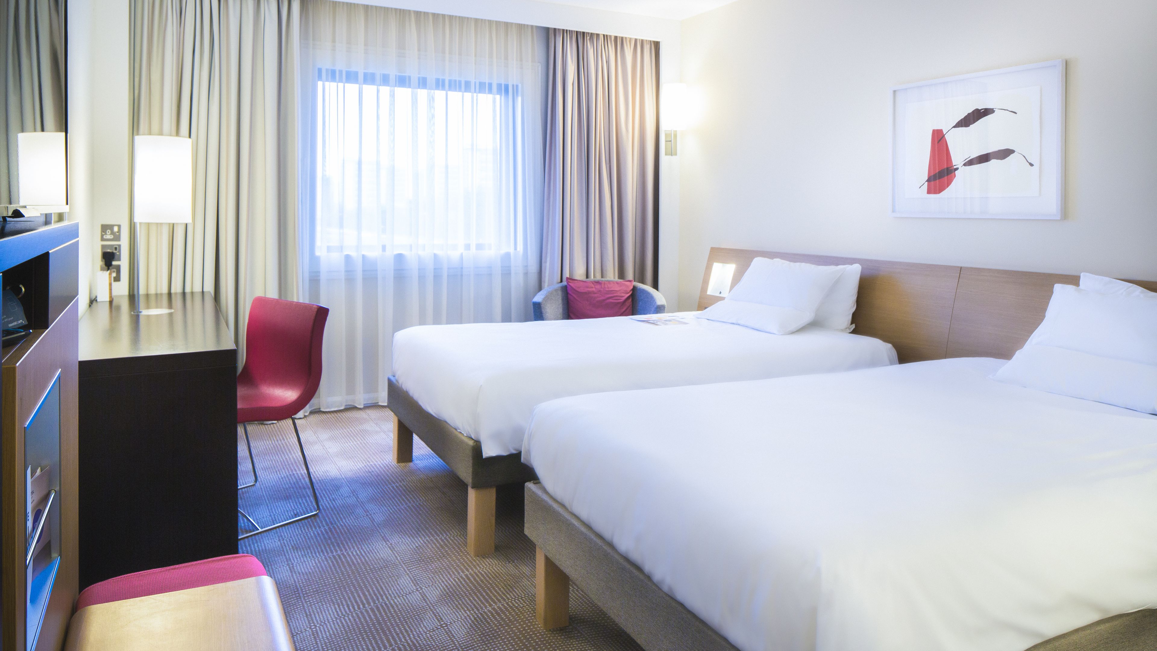 A hotel room with two beds, a desk, a chair, and a window with curtains.