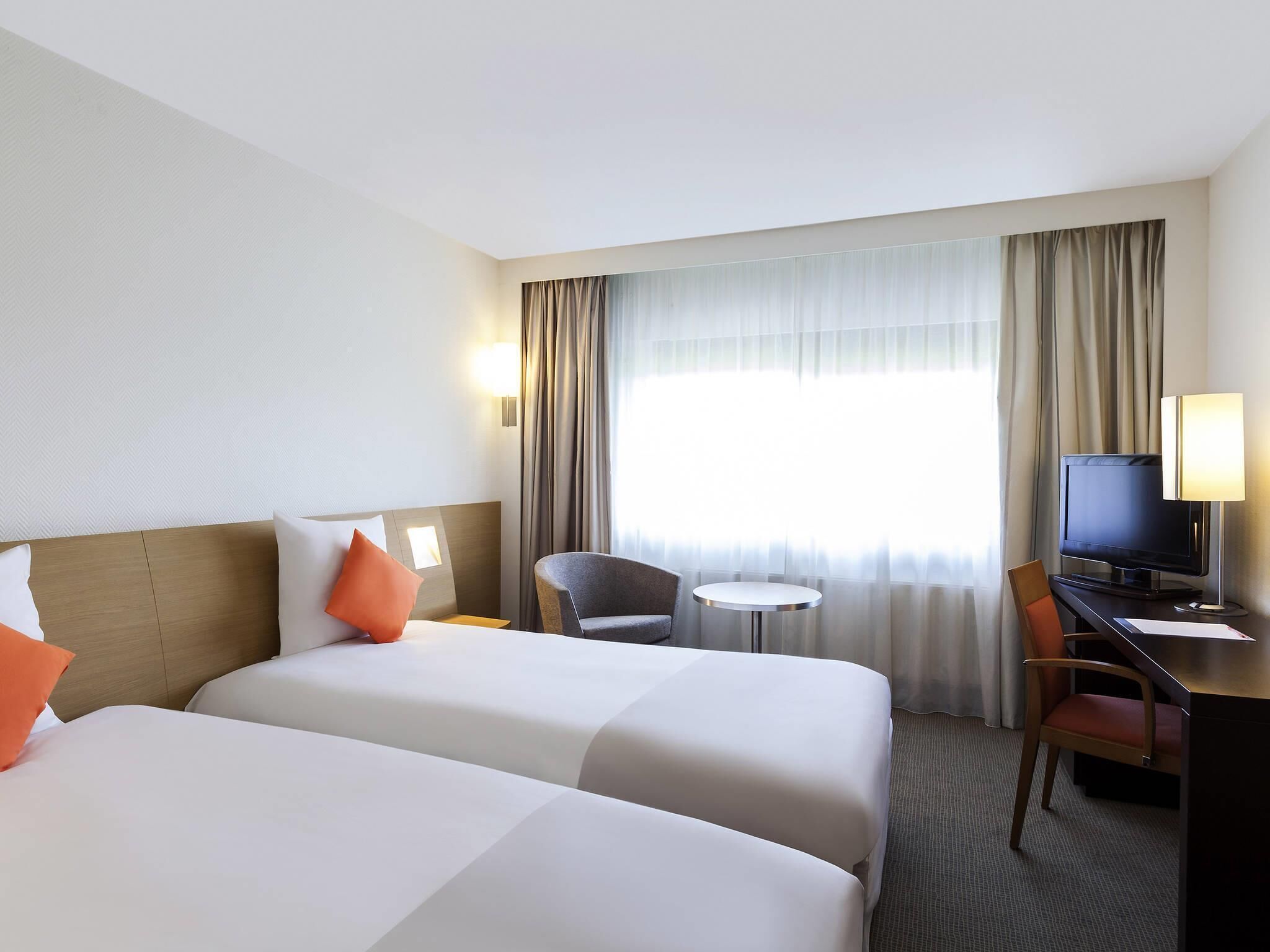 A hotel room with two beds, a desk with a TV, a chair, a small table, and a window with curtains.