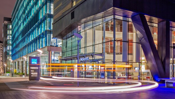 A modern hotel with a glass facade, a Novotel sign, and a light trail indicating movement. - Novotel London Paddington (London)