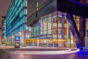 A modern hotel with a glass facade, a Novotel sign, and a light trail indicating movement. - Novotel London Paddington (London)