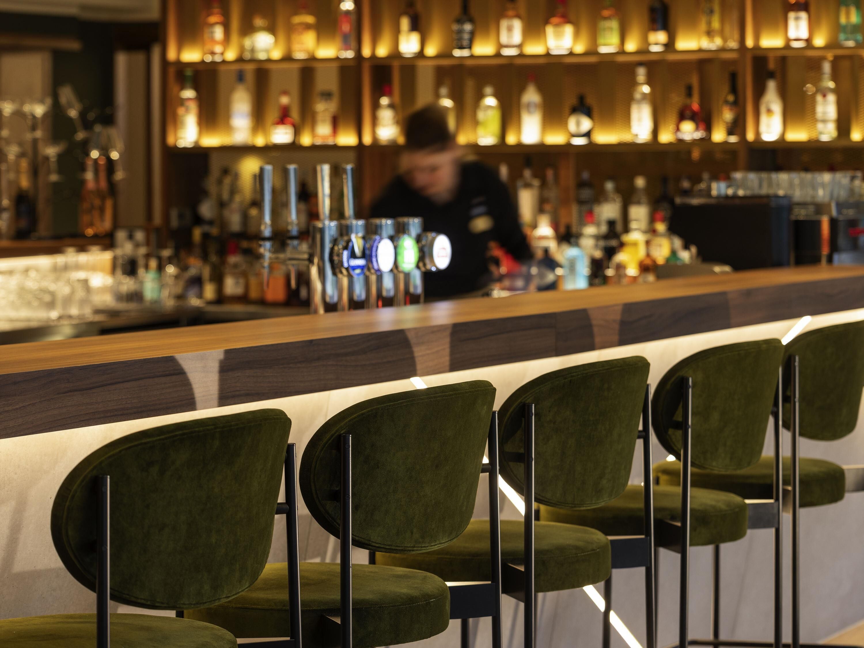 Una zona de bar con sillas tapizadas de verde y un bartender detrás de la barra.
